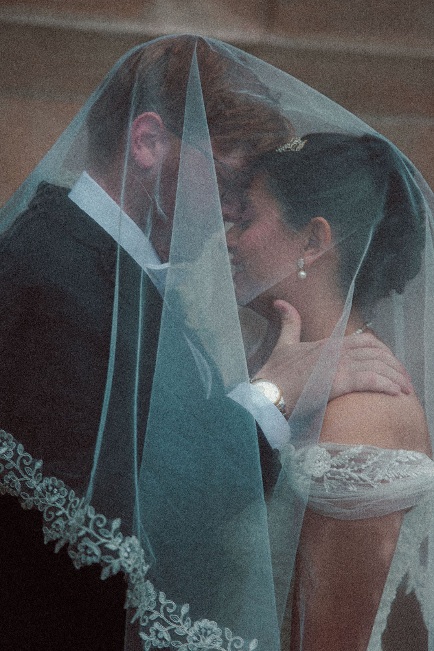 Bride and groom kissing under a lace-edged wedding veil outdoors.
