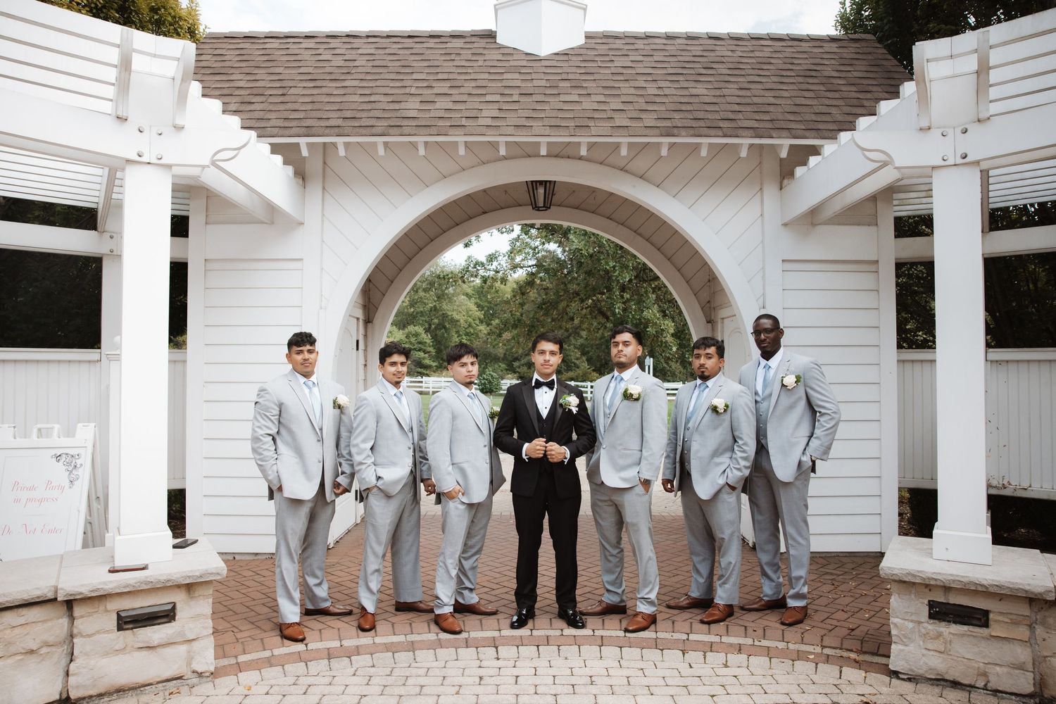 Groom surrounded by groomsmen in light gray suits under a white pergola arch.