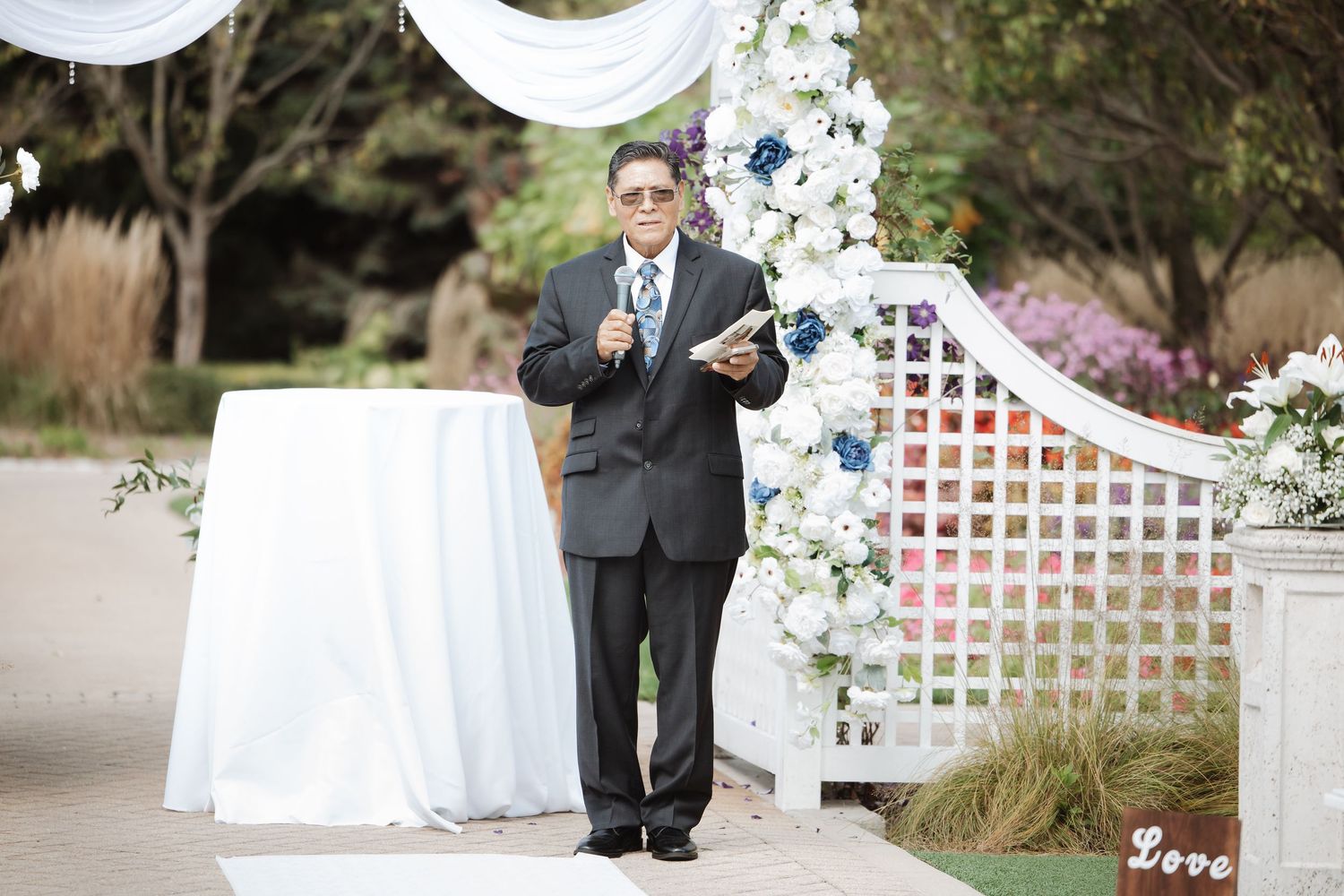 Officiant speaks at outdoor wedding ceremony near floral arch with white flowers and greenery.