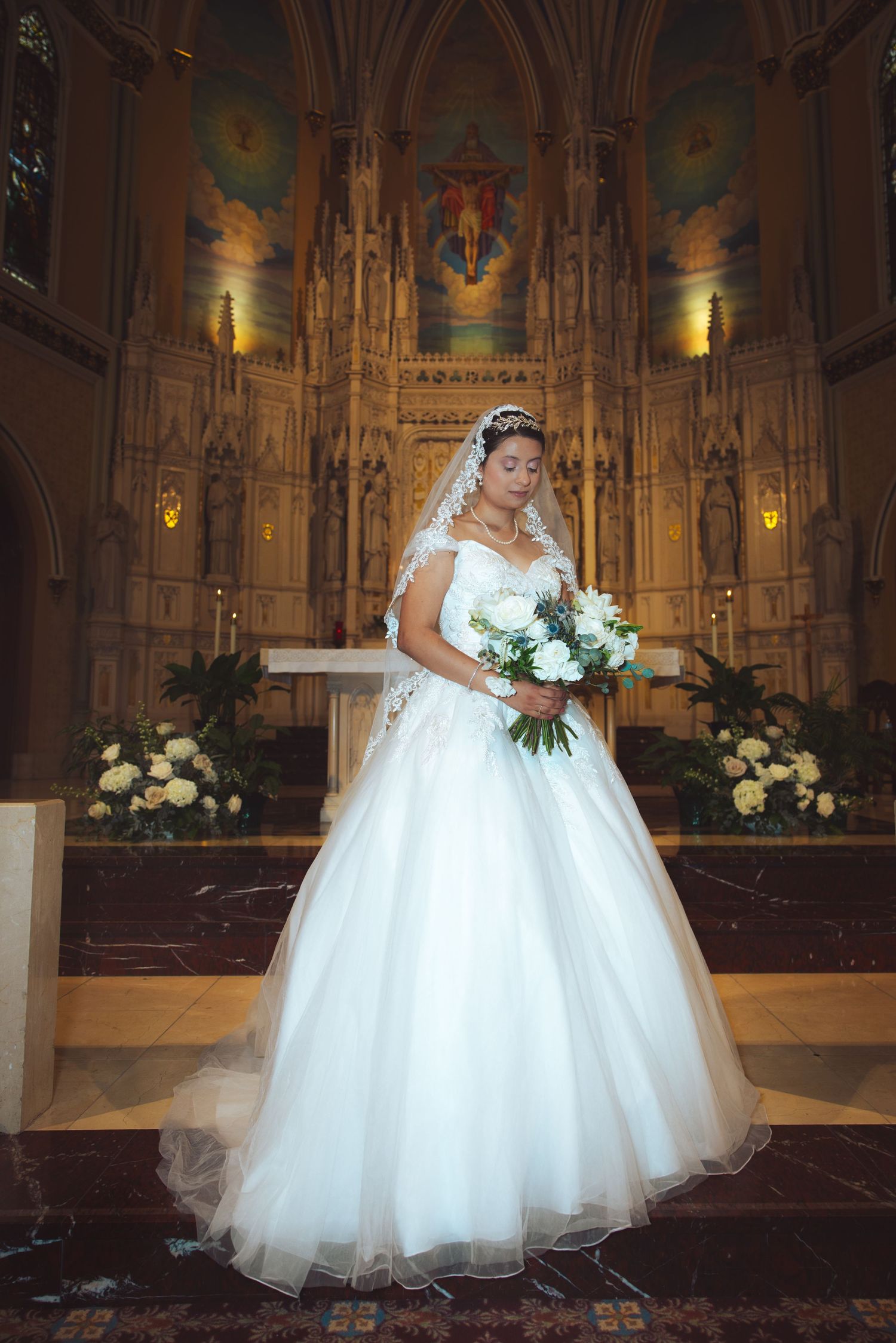 Elegant bride in white ball gown holding white bouquet standing before grand Gothic church altar.