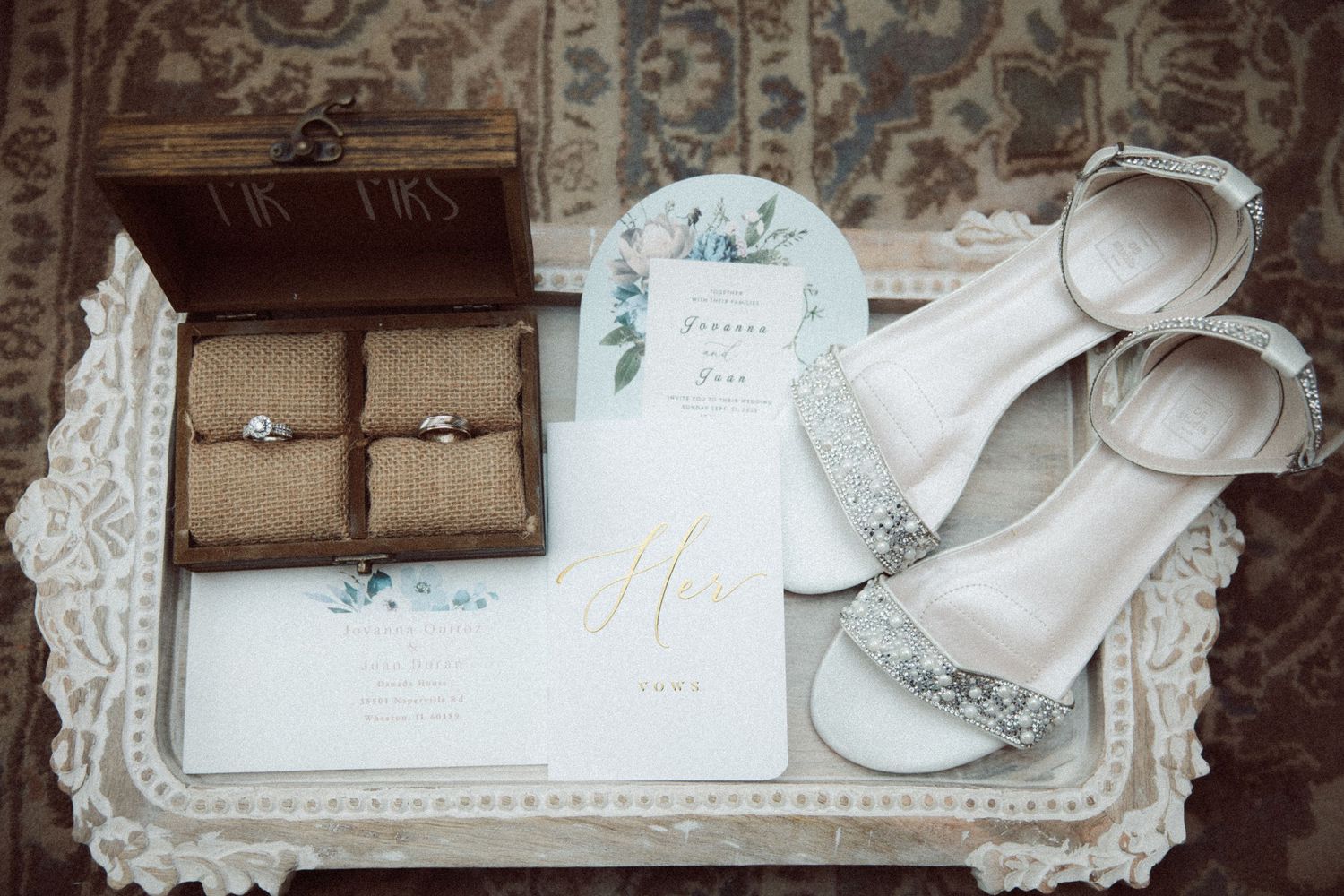 Wedding detail flat lay with diamond rings in wooden box, glittery heels, and floral stationery on tray.