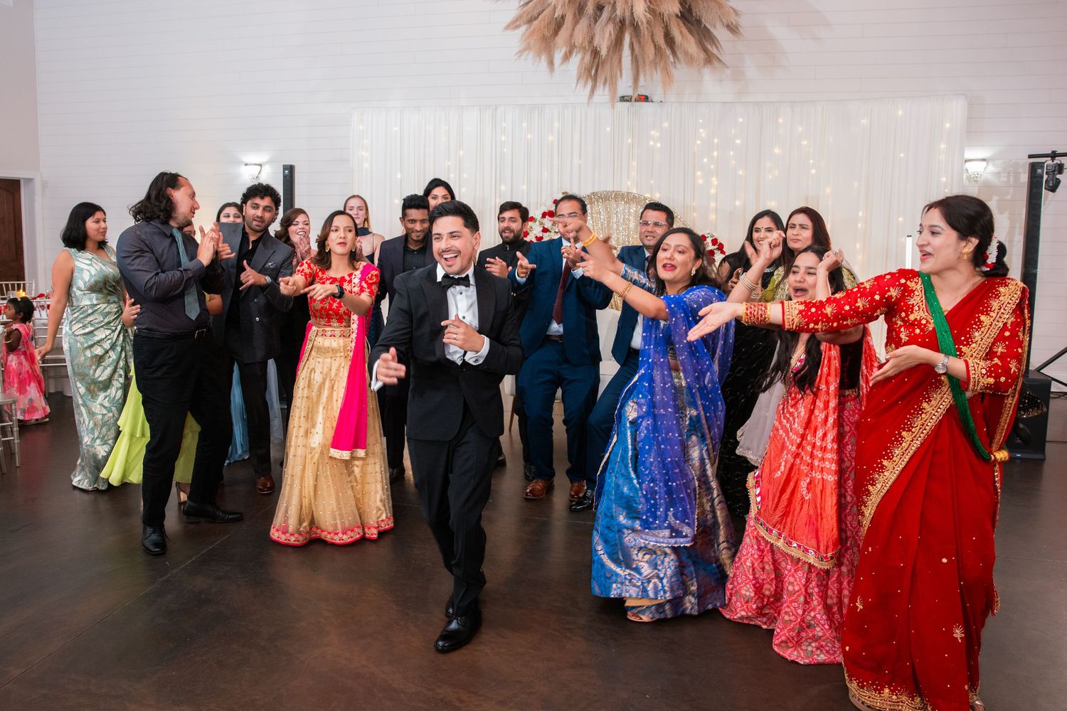 Groom in tuxedo dances with enthusiastic wedding guests in colorful traditional Indian attire at reception.