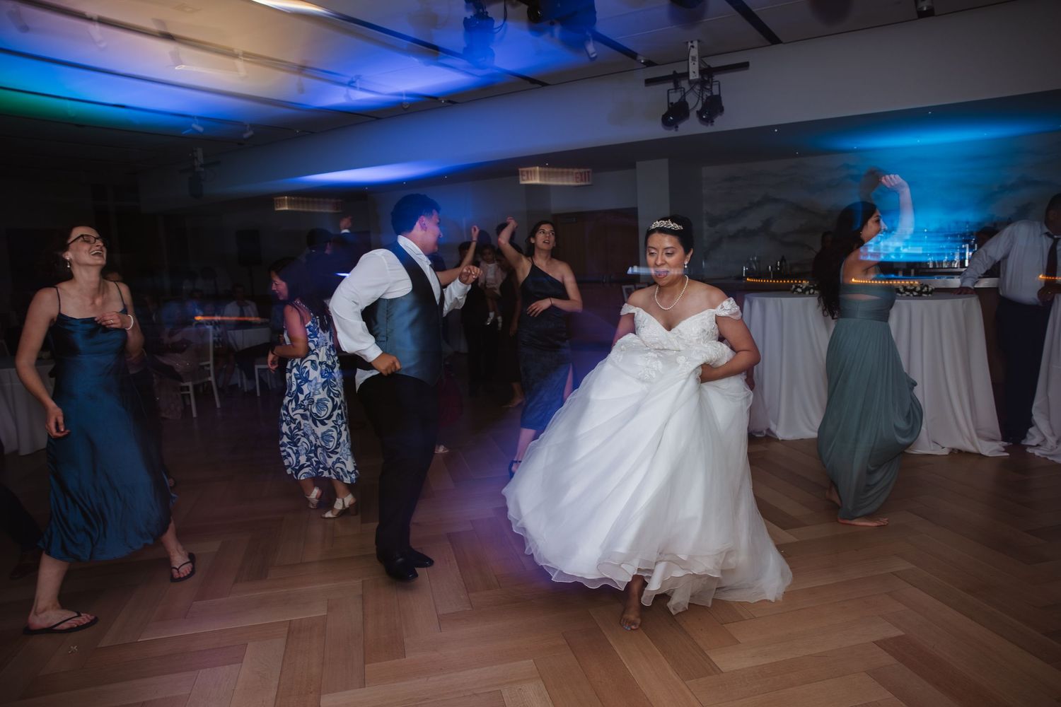 Bride in white ball gown dancing with guests at wedding reception with blue ambient lighting.