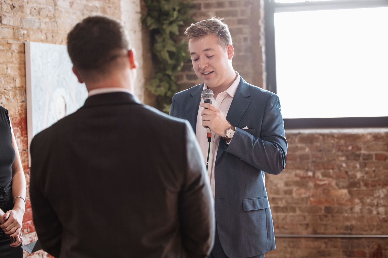 Man in blue suit speaking into microphone at indoor wedding ceremony with exposed brick wall backdrop.