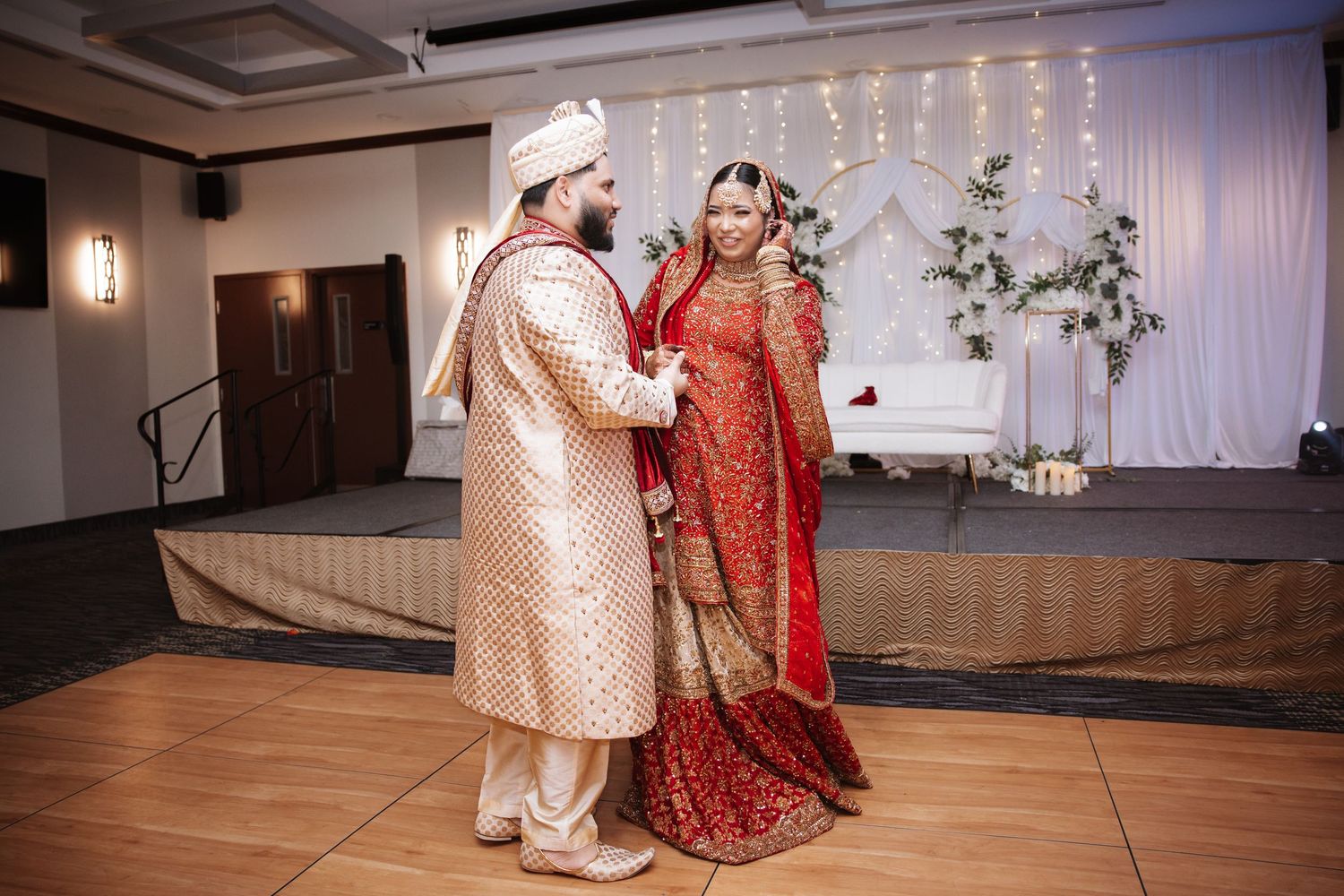 Indian bride and groom in traditional red and gold wedding attire sharing first dance on ballroom floor.