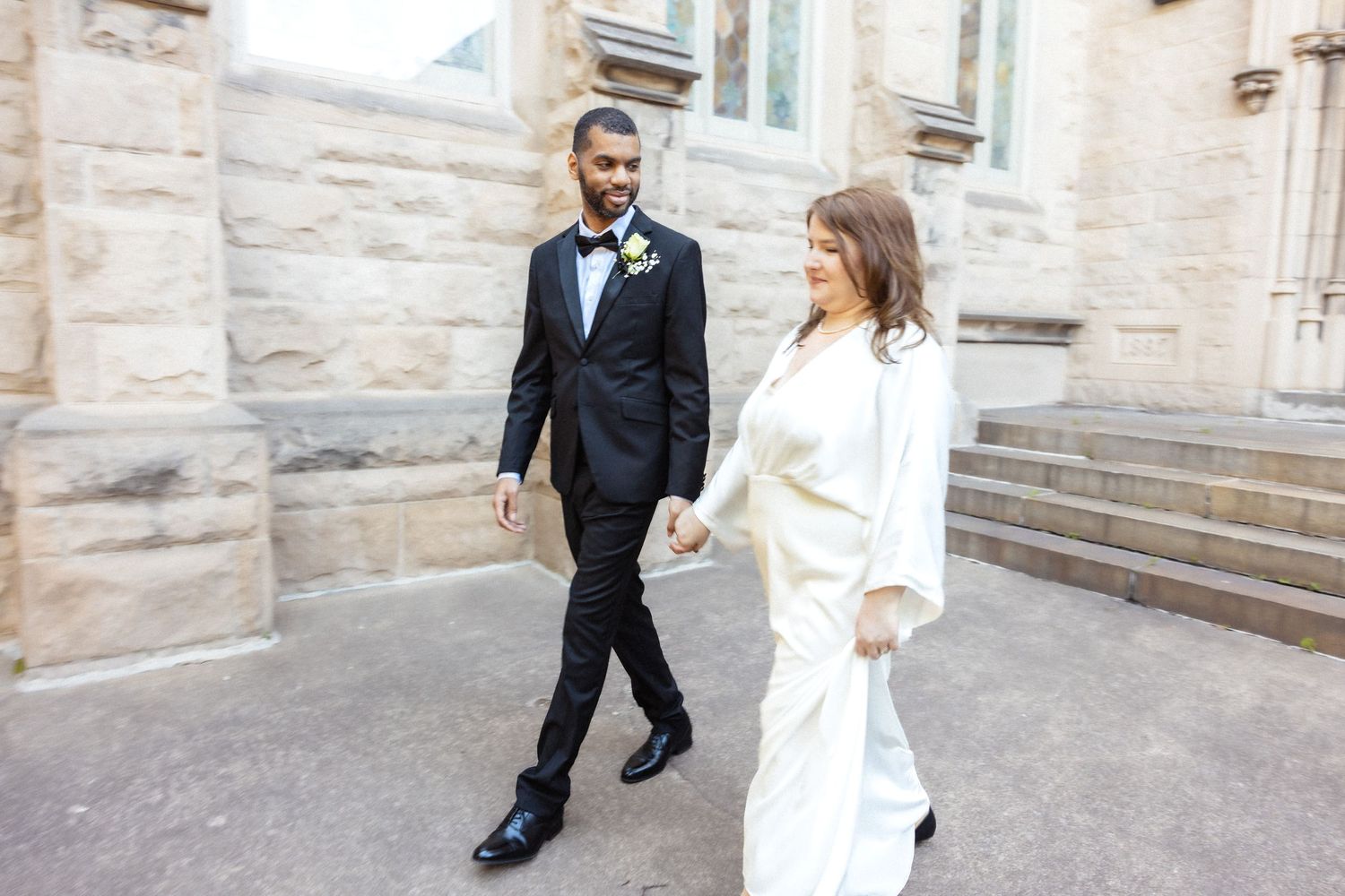 Elegant couple walking hand in hand outside a stone church on their wedding day.