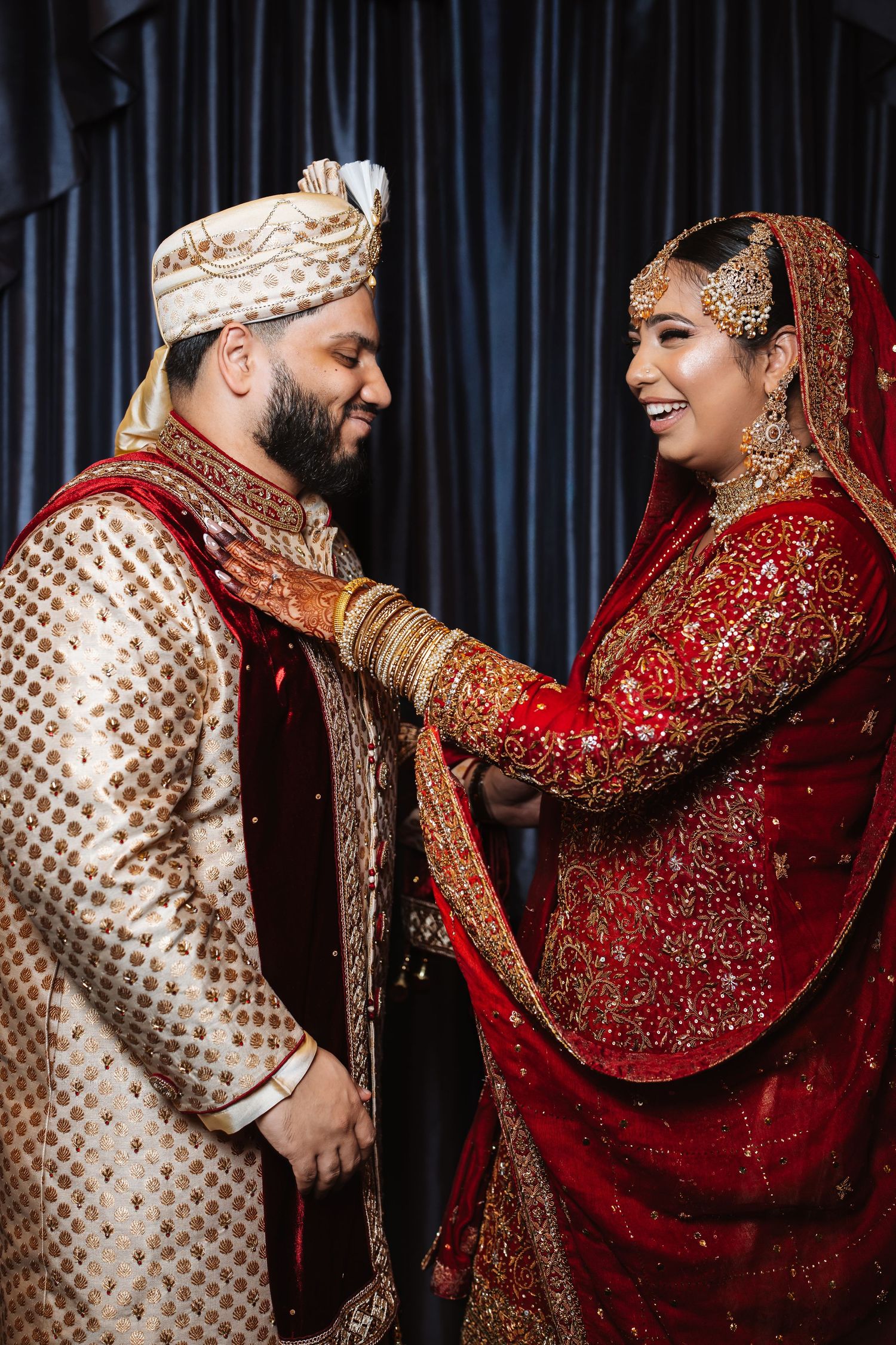 South Asian bride in red lehenga laughs with groom in gold sherwani at traditional wedding.