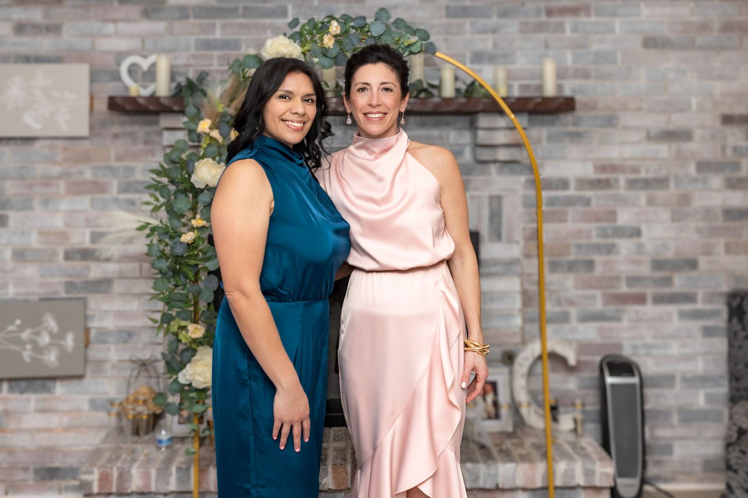 Two women in elegant formal attire smiling together at an event with golden arch backdrop.