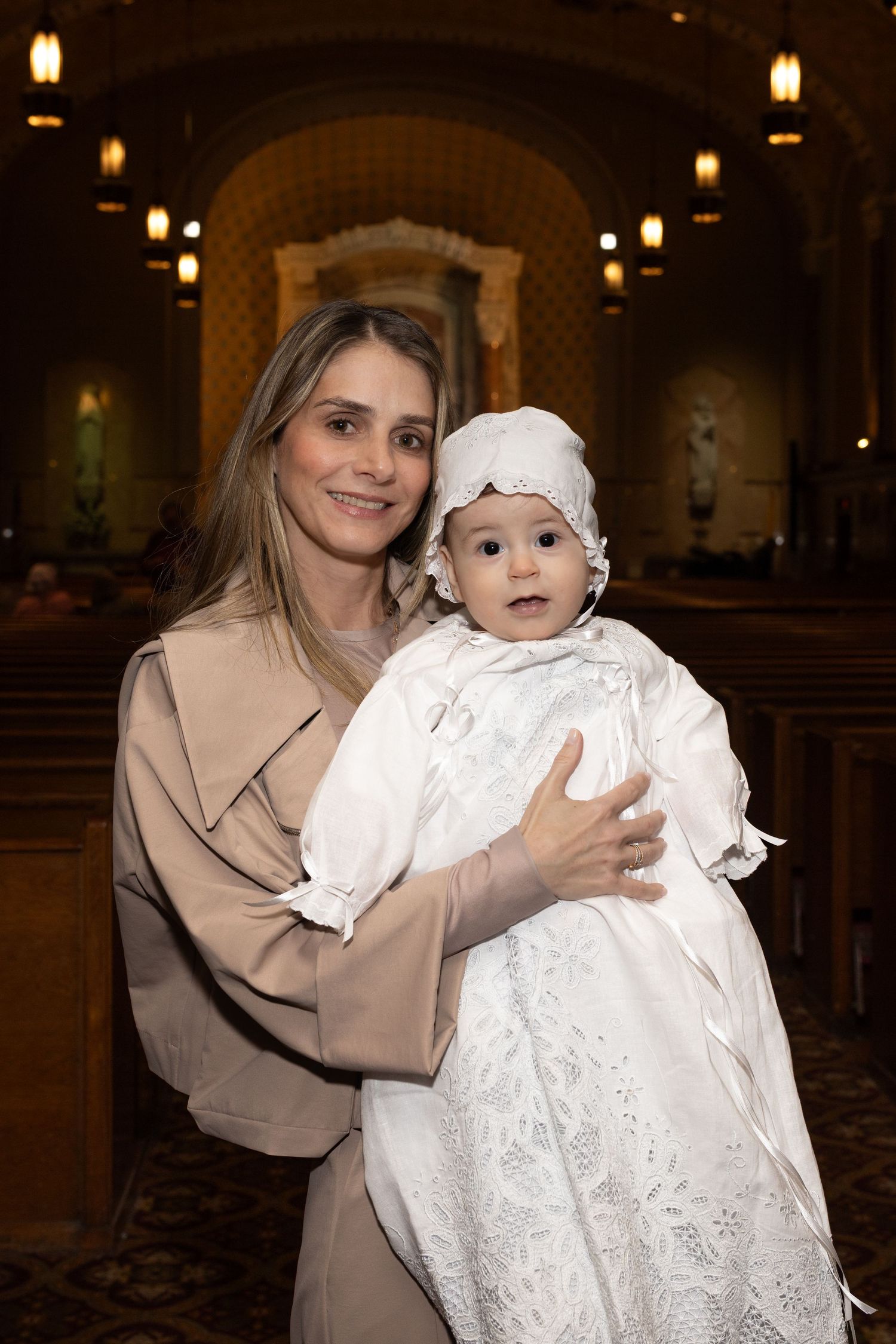 Woman in beige coat holds infant in white christening gown and bonnet inside a church.