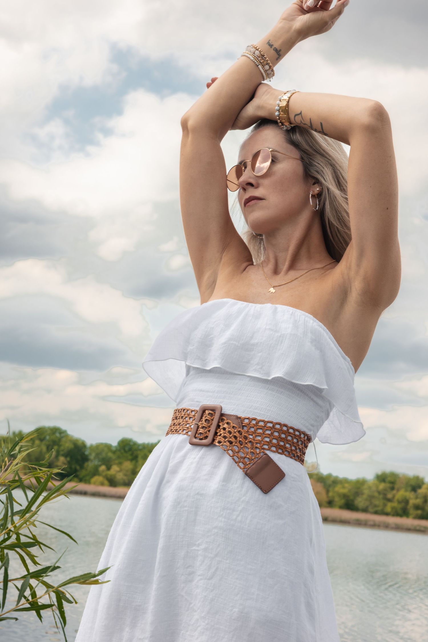 Woman in a white strapless ruffled dress with a brown belt posing outdoors near a lake.