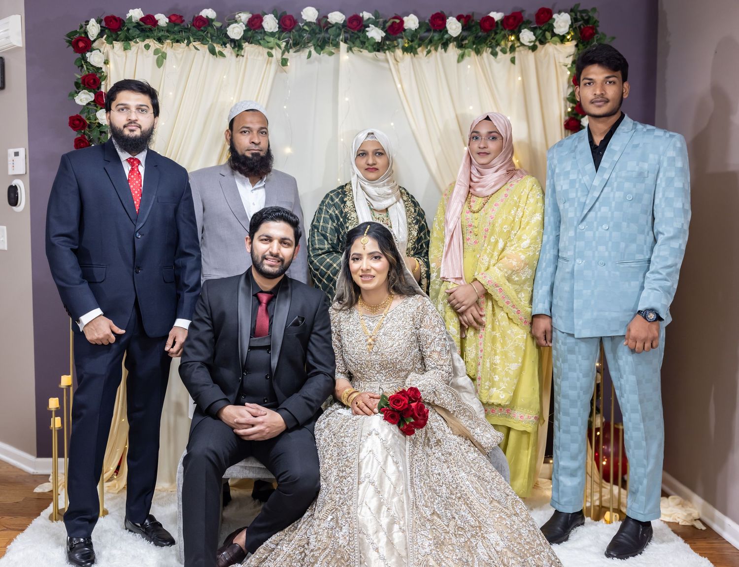 Wedding party poses with bride and groom in elegant attire before floral backdrop at South Asian ceremony.