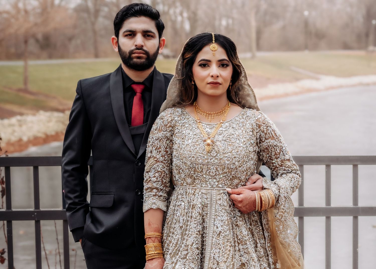Pakistani couple pose together outdoors, bride in ornate silver-gold lehenga, groom in black suit with red tie.