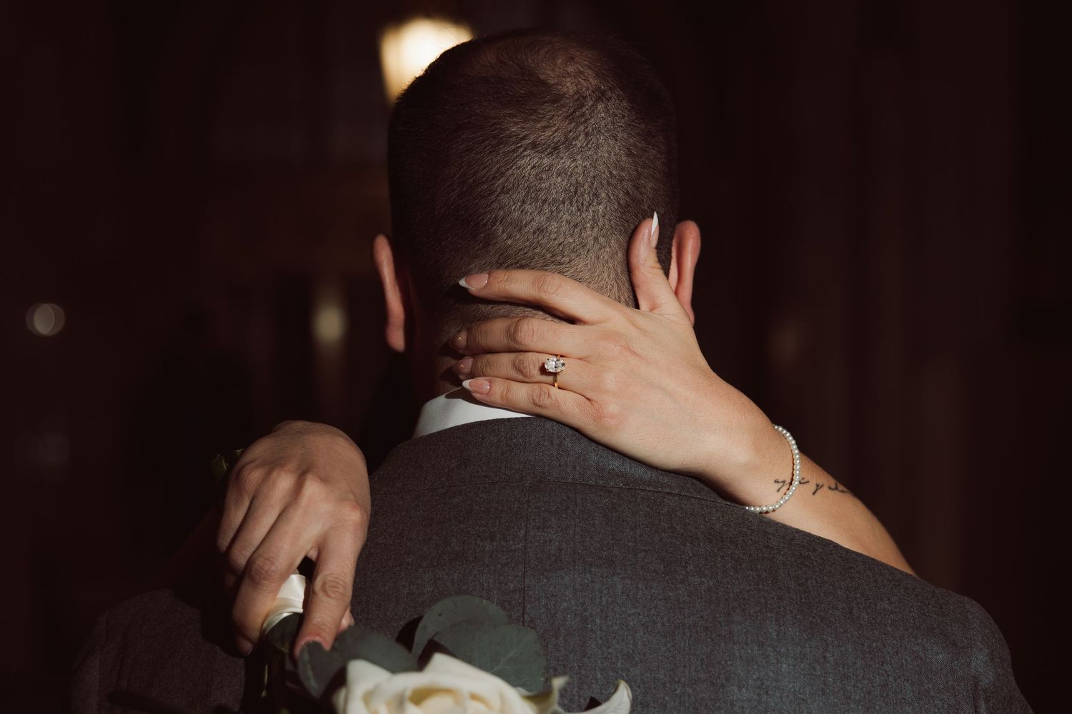 Bride placing hand on grooms neck holding white bouquet from behind in dark setting.