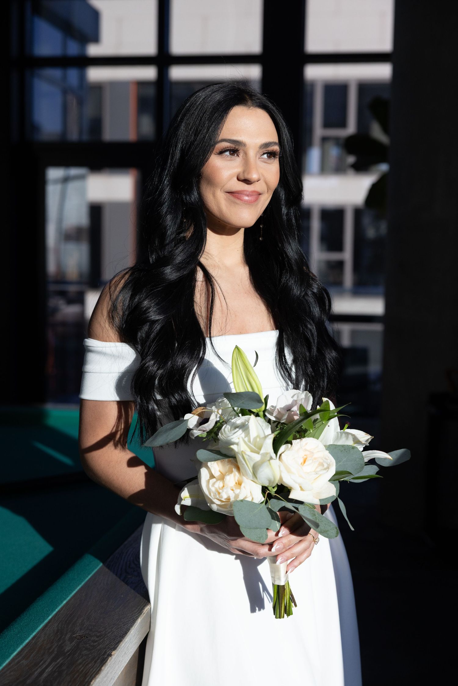 Beautiful dark-haired bride in white off-shoulder gown holding white rose bouquet outdoors.