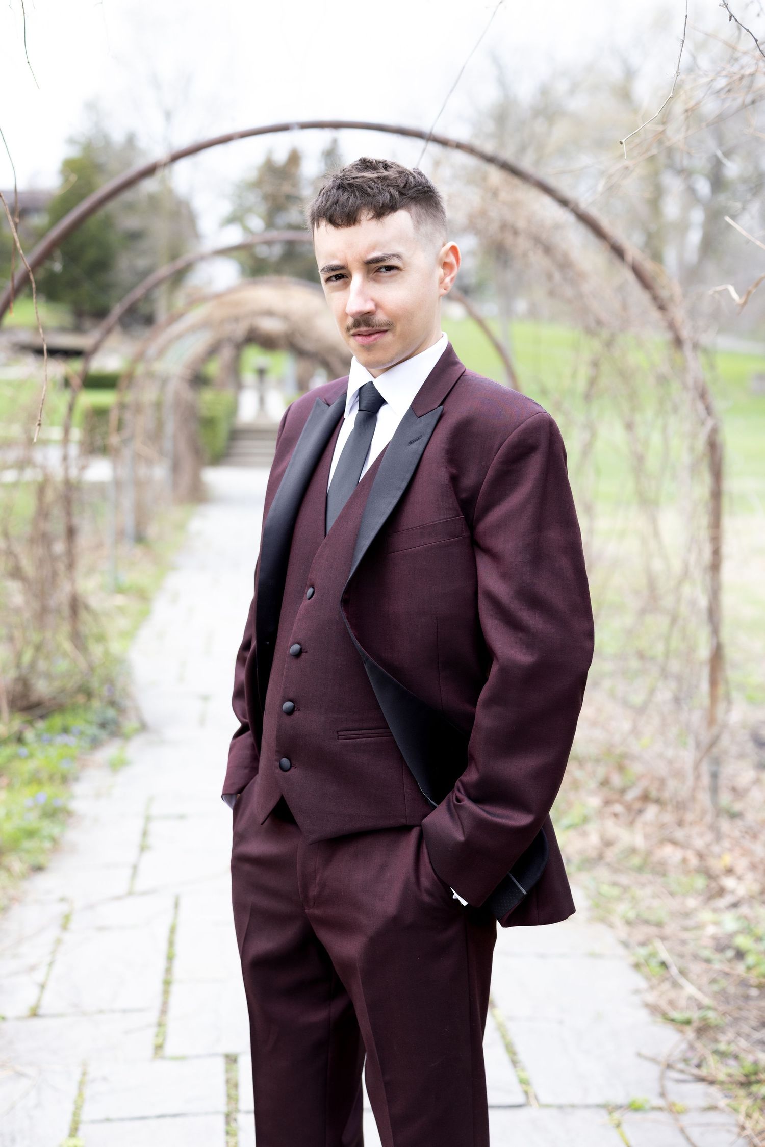 Man in burgundy three-piece tuxedo with mustache poses on garden path under metal arch.