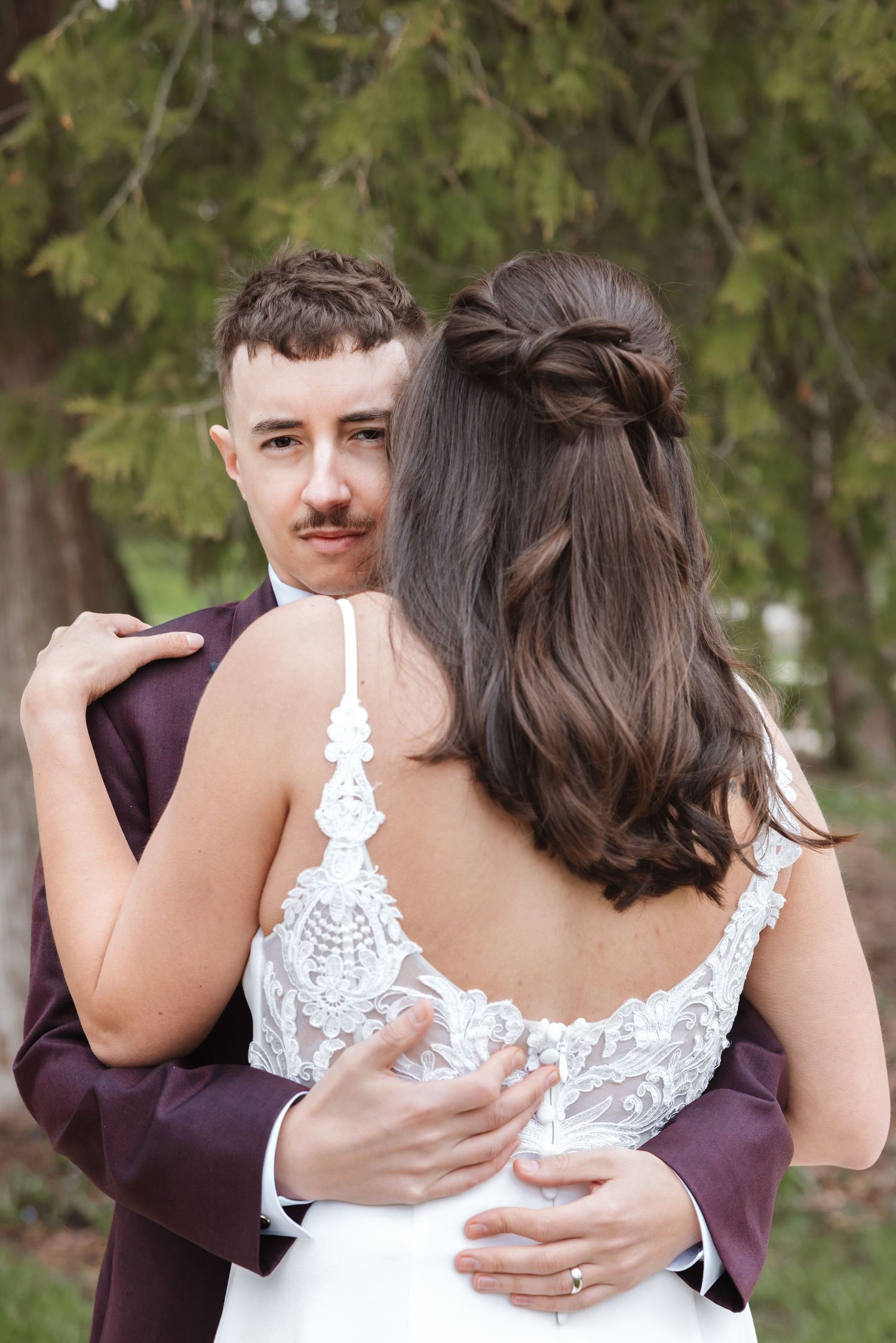 Bride and groom embracing outdoors, groom in burgundy suit looking at camera, bride in lace backless wedding dress.