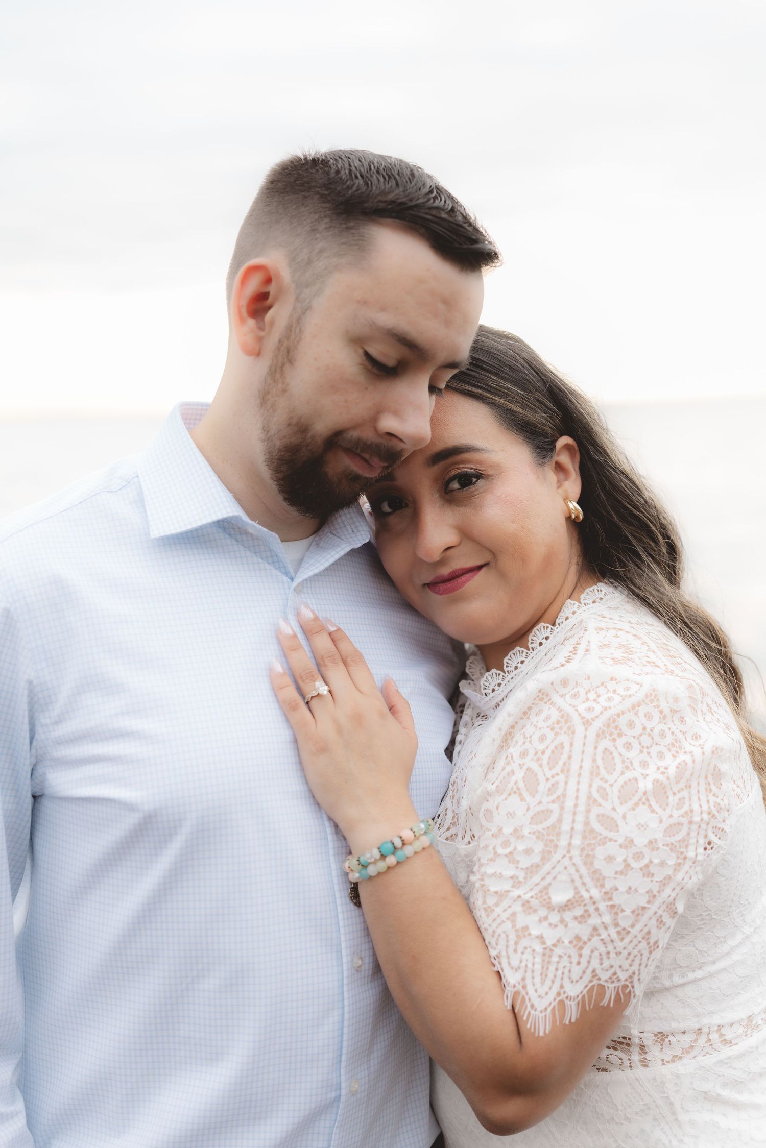 Couple embracing tenderly outdoors, woman in white lace dress leaning on mans chest, wearing engagement ring.