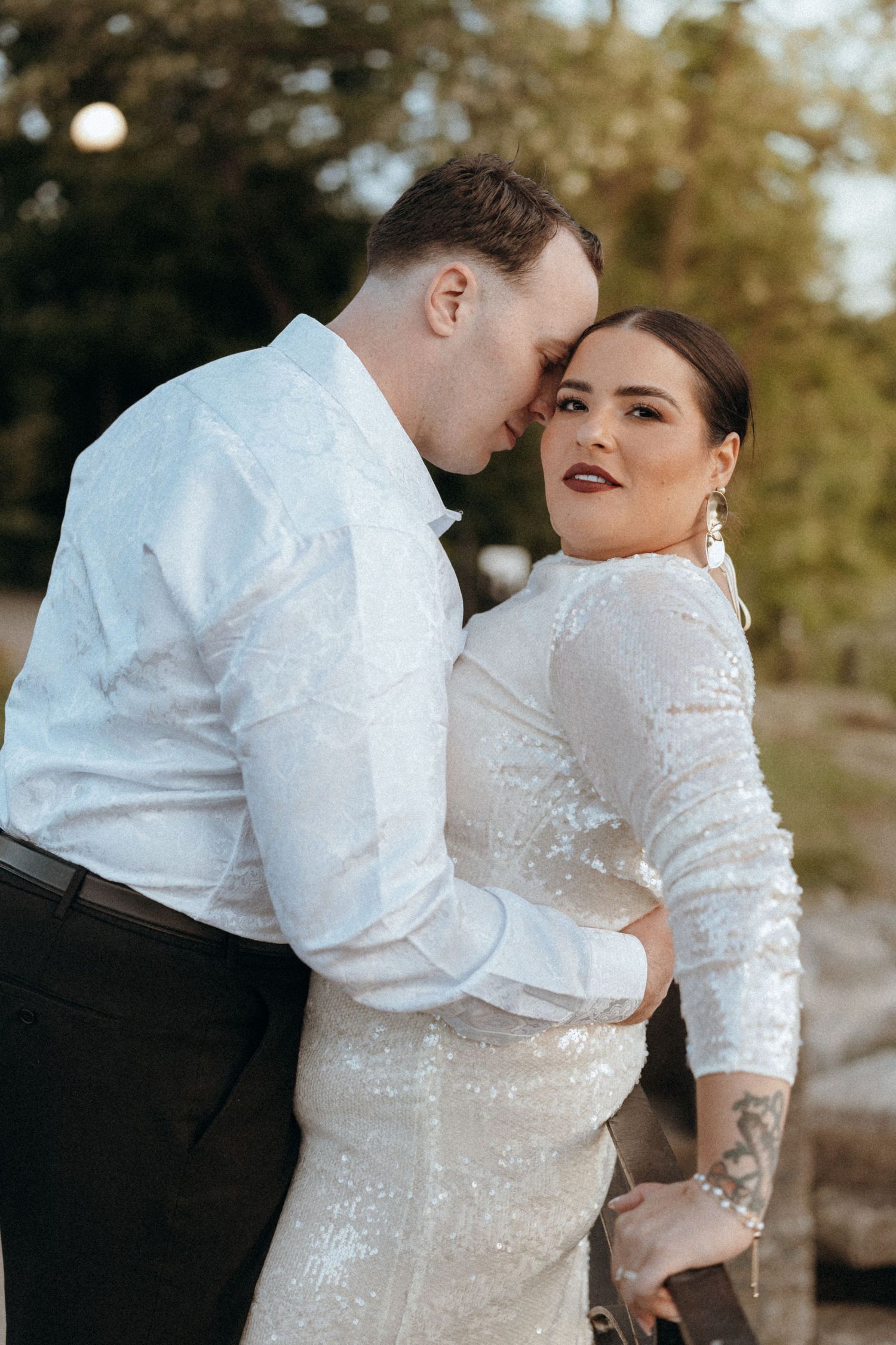 Groom in white shirt embraces bride in sequin wedding dress during golden hour outdoor portrait.