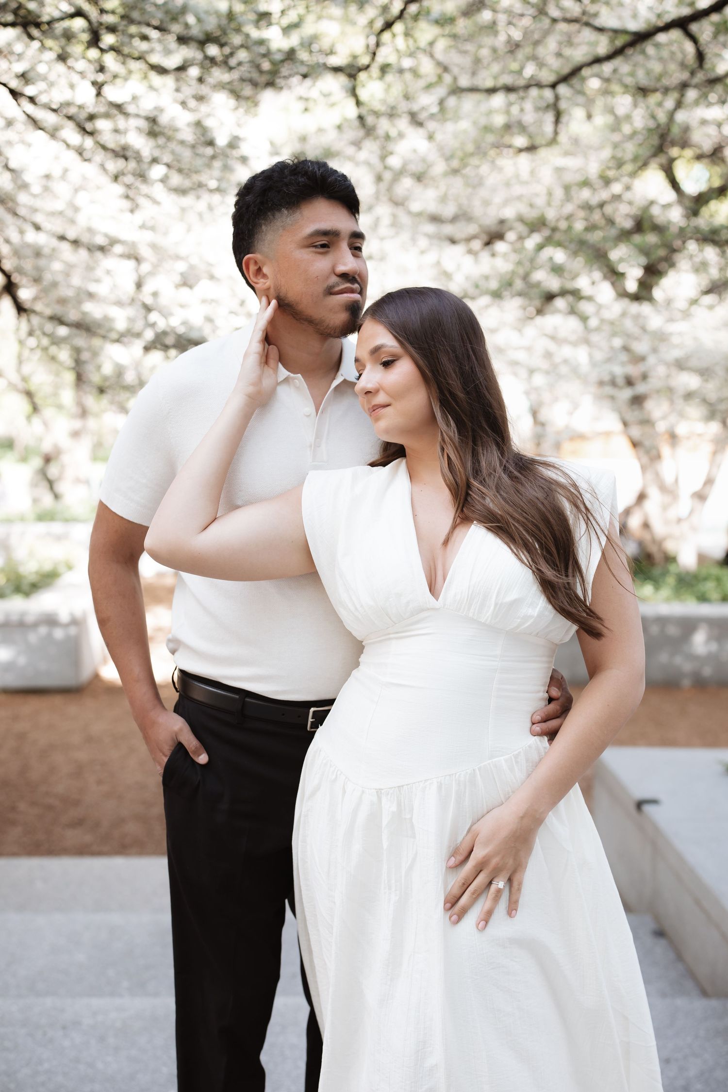 Couple embracing outdoors under blooming trees, woman in white dress leaning on man in white polo and black pants.