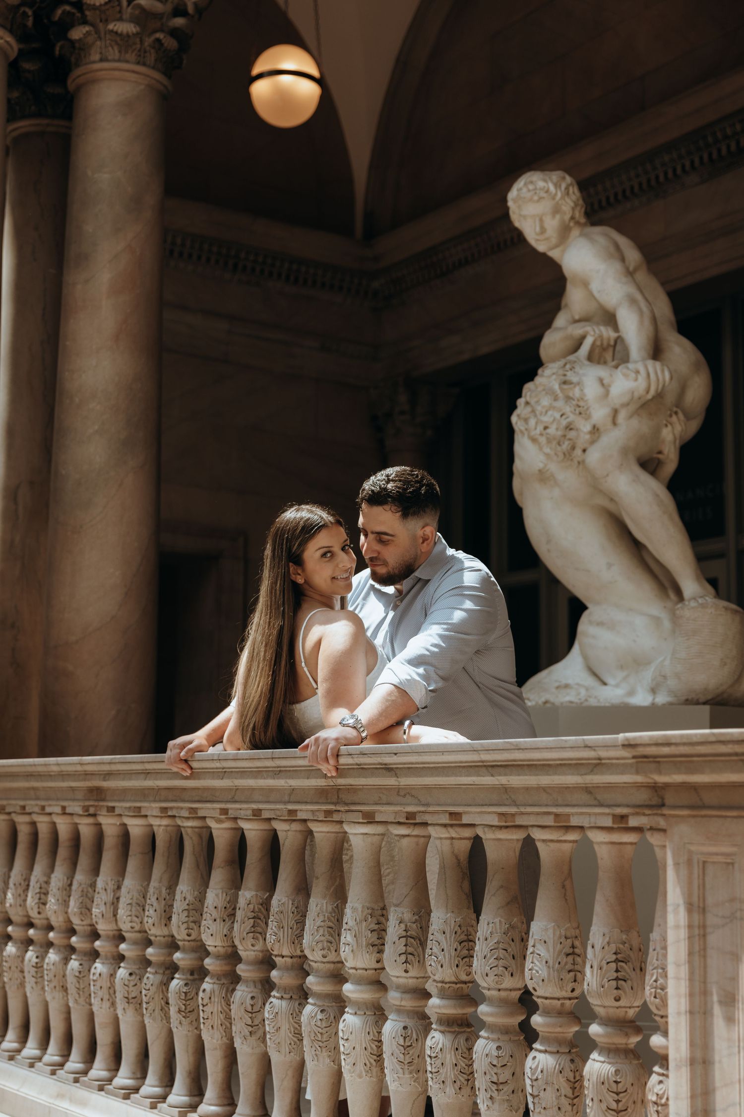 Elegant couple leaning on ornate marble balustrade near classical sculpture in a grand architectural interior.