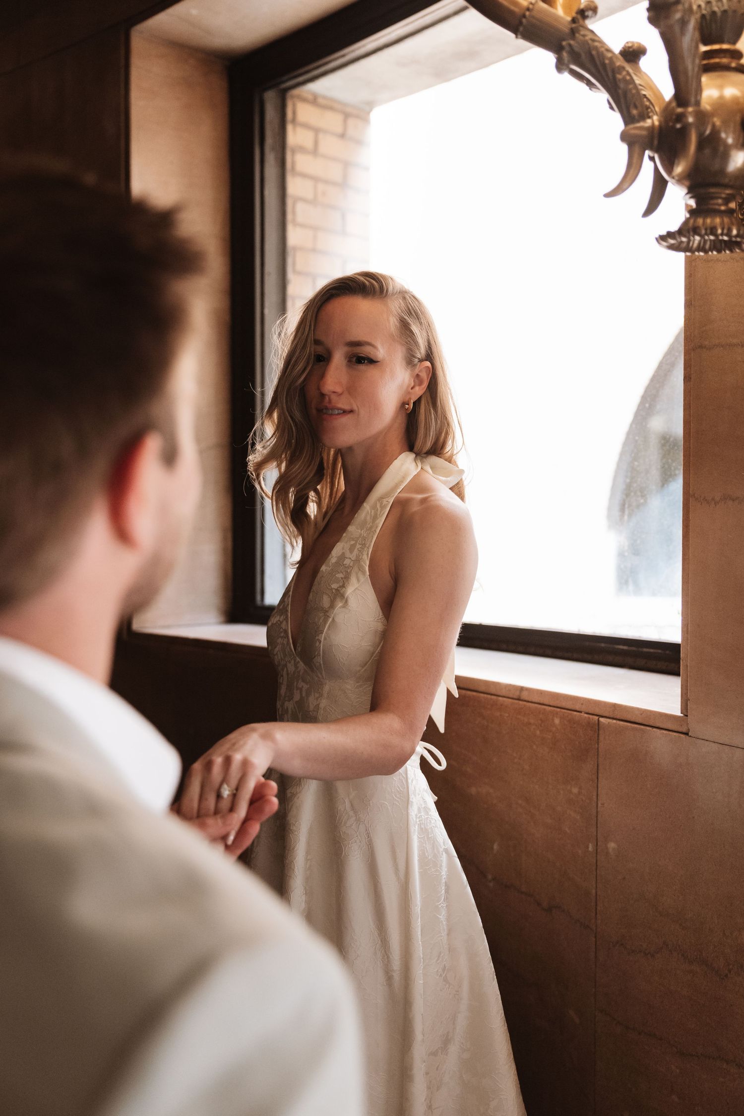 Bride in white halter dress holds groom's hand near sunlit window in romantic wedding portrait.