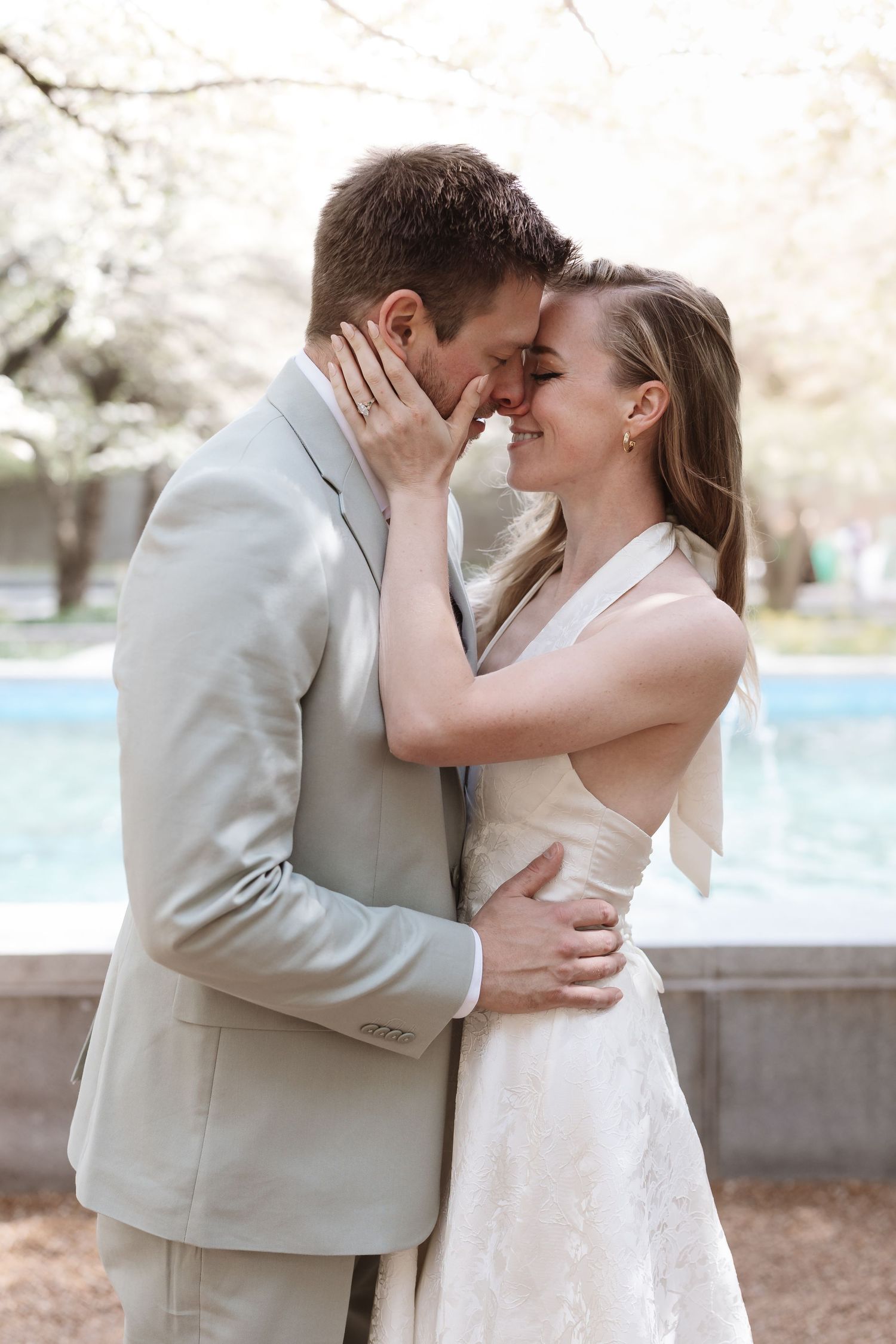 Smiling bride and groom nearly kissing outdoors by fountain with cherry blossoms.