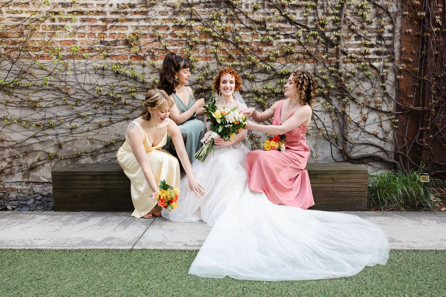 Bride in a white tulle gown sits with three bridesmaids in colorful dresses against an ivy-covered brick wall.