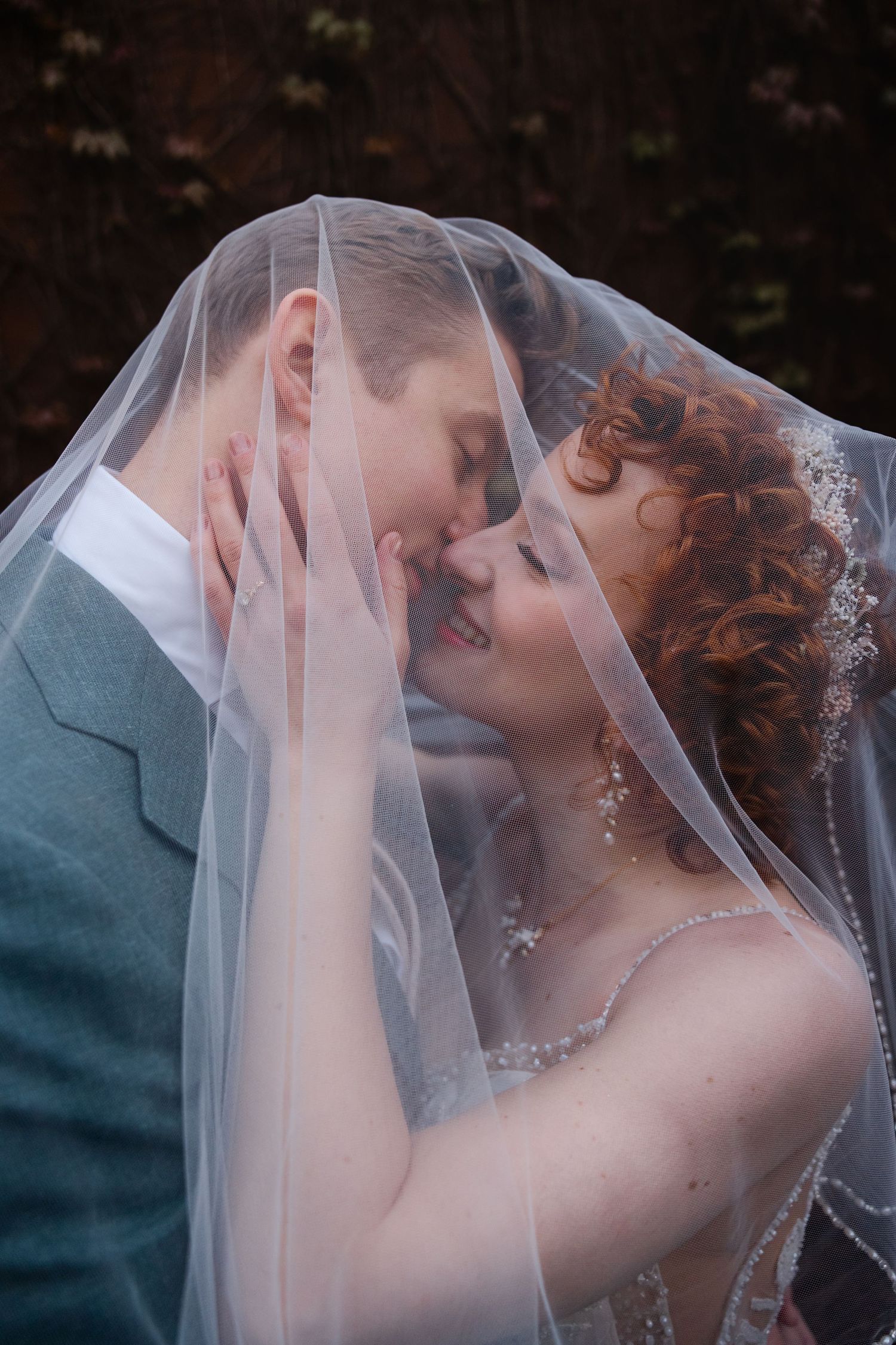 Bride and groom sharing a romantic kiss under a flowing white wedding veil outdoors.
