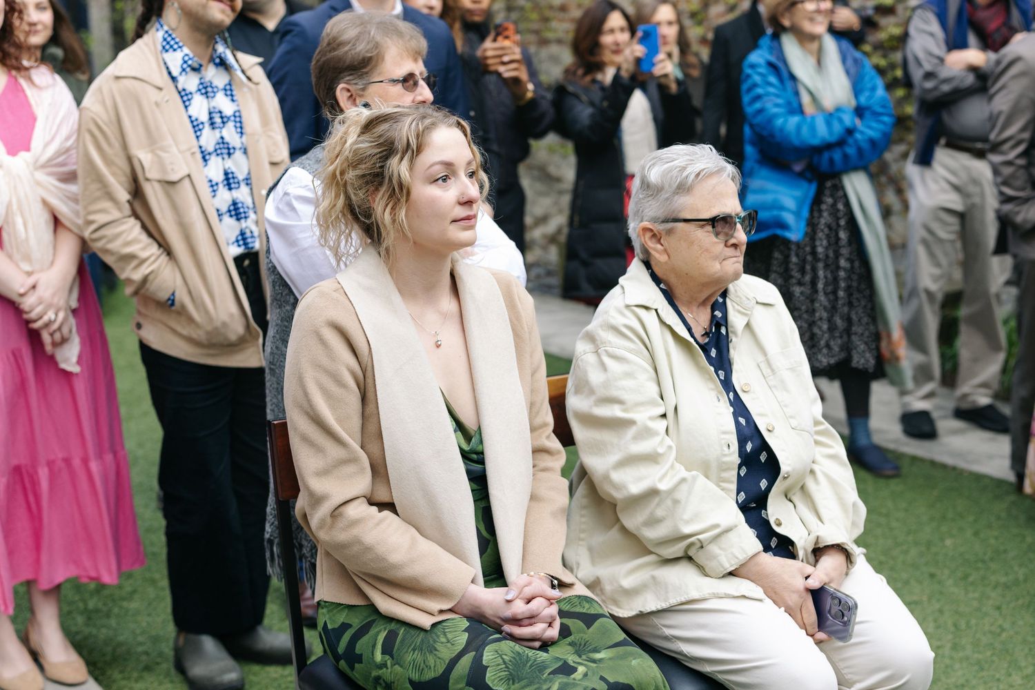Two women seated outdoors at a wedding ceremony, surrounded by standing guests in a garden setting.