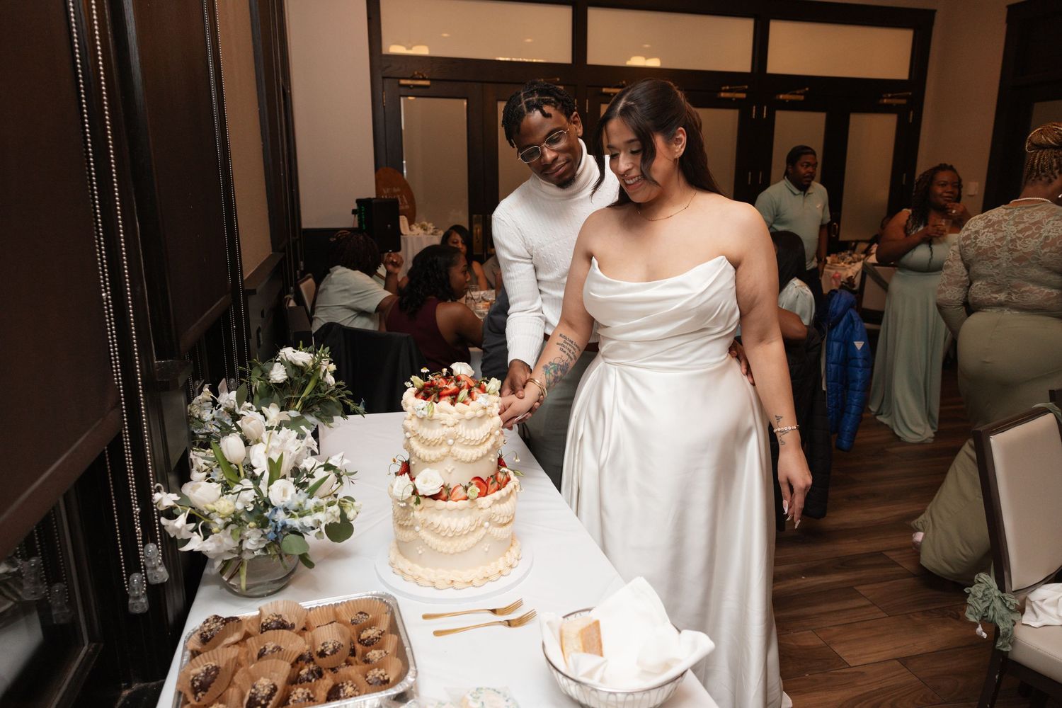 Bride and groom cut elegant naked wedding cake at reception surrounded by guests.
