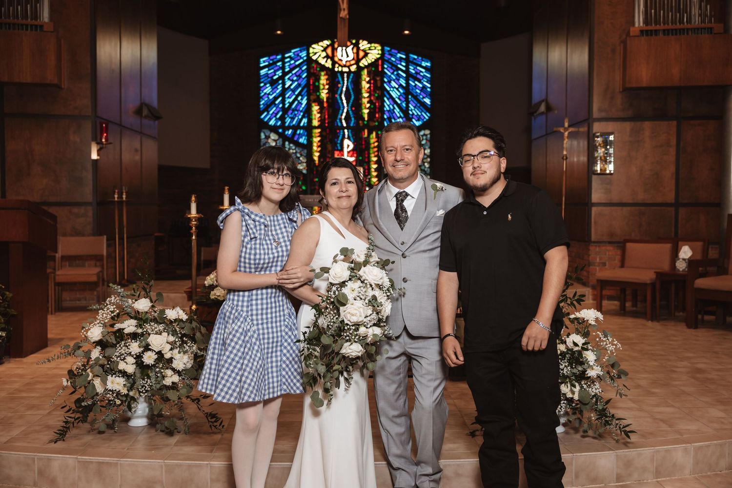 Wedding party of four poses at church altar with floral arrangements and stained glass window backdrop.
