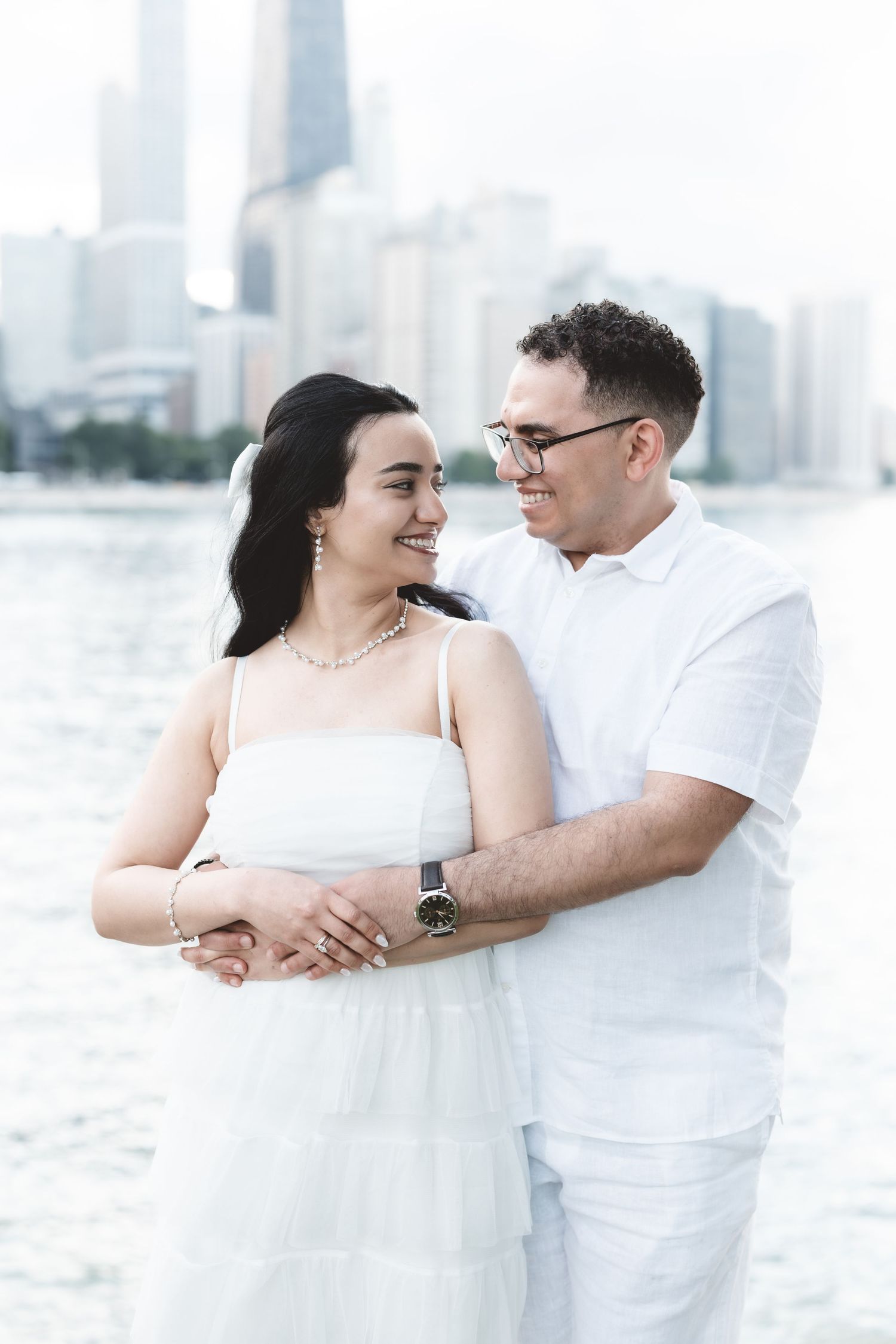 Couple in matching white outfits embracing and smiling by a city waterfront.
