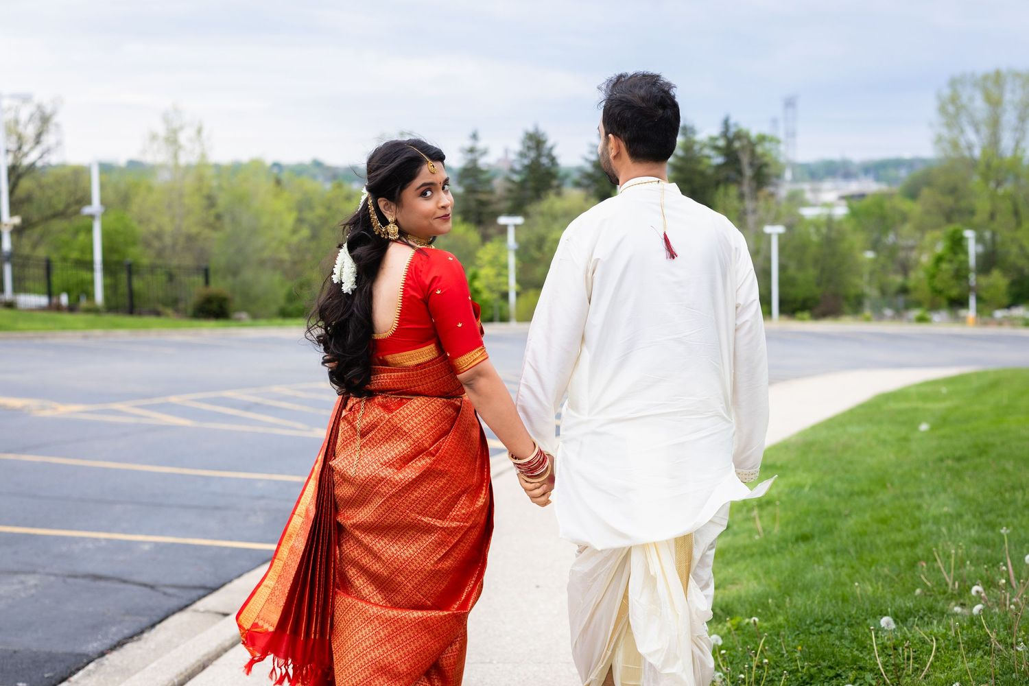 Indian couple in traditional attire holding hands while walking on a sidewalk, woman in red saree, man in white kurta.
