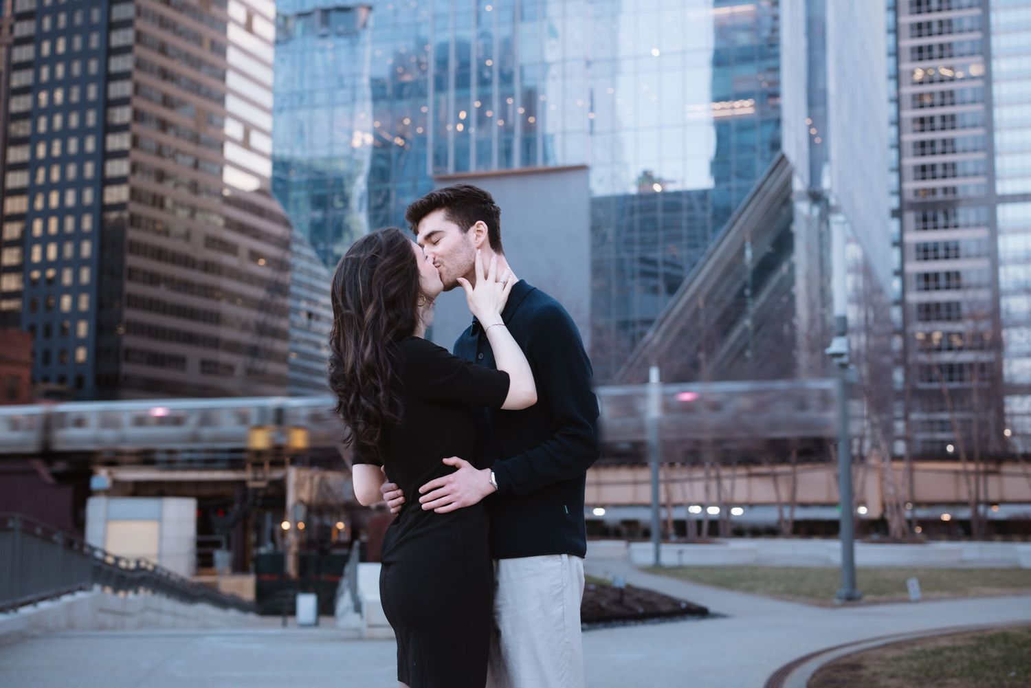 Couple sharing a kiss outdoors with Chicago city buildings and elevated train in background.