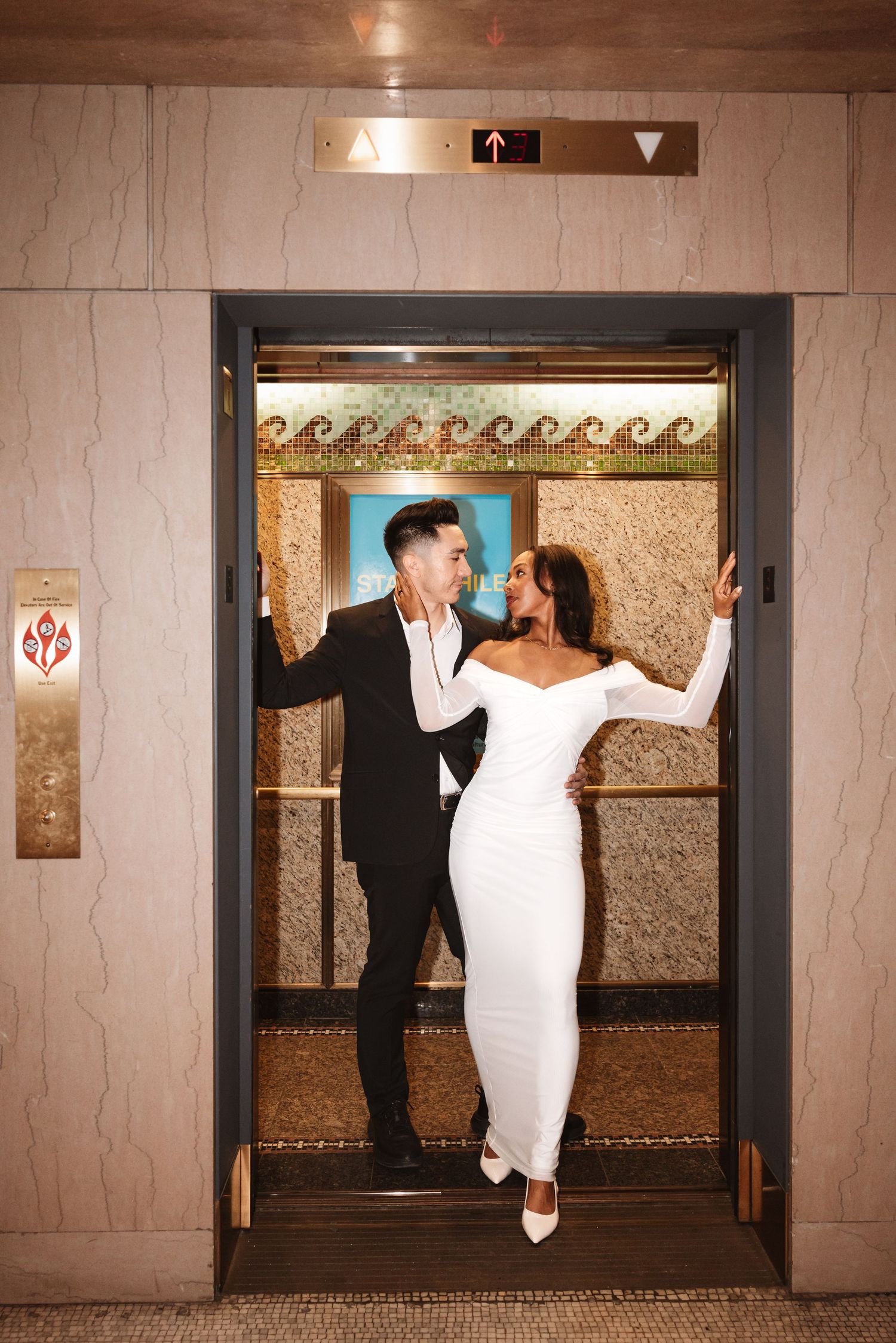 Bride and groom smiling at each other while standing in an open elevator doorway.