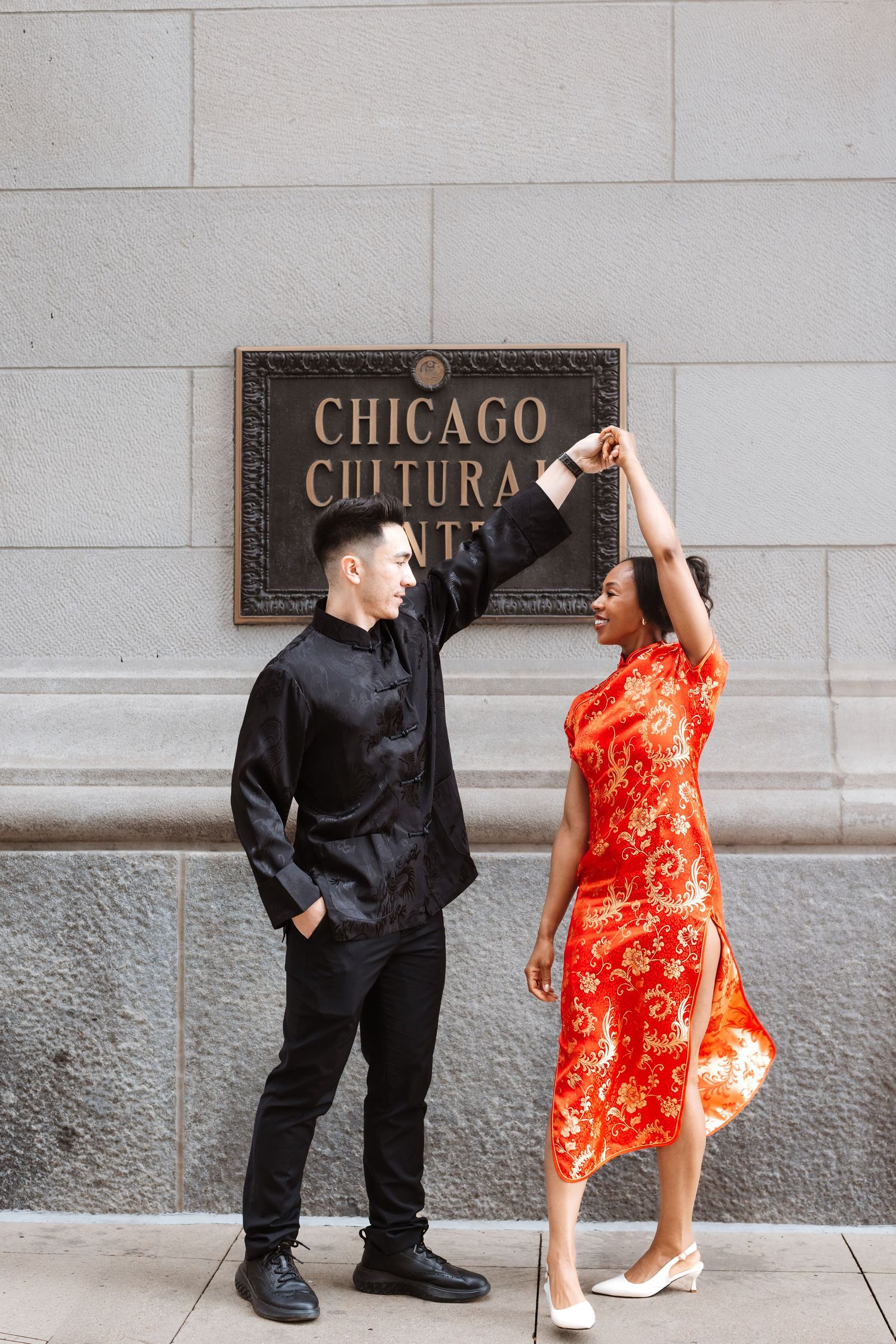 Man in black tang suit twirling woman in red floral qipao outside Chicago Cultural Center.