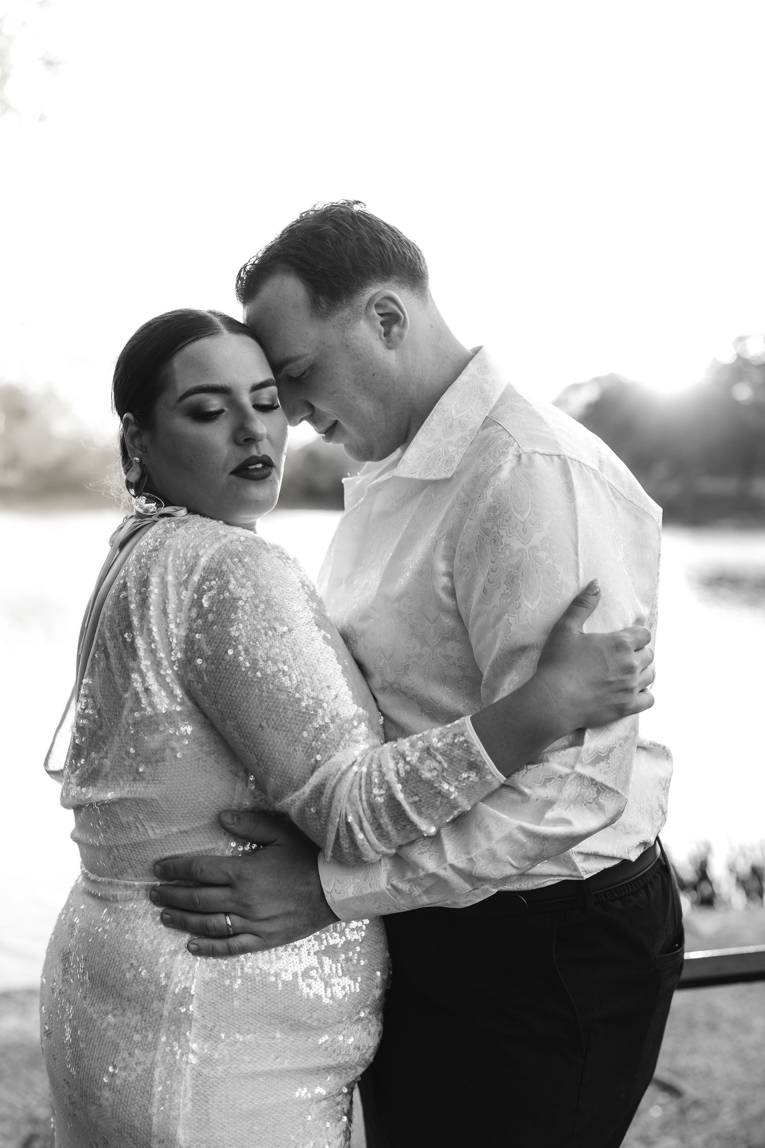 A black and white romantic portrait of a bride and groom embracing tenderly near a sunlit waterfront.