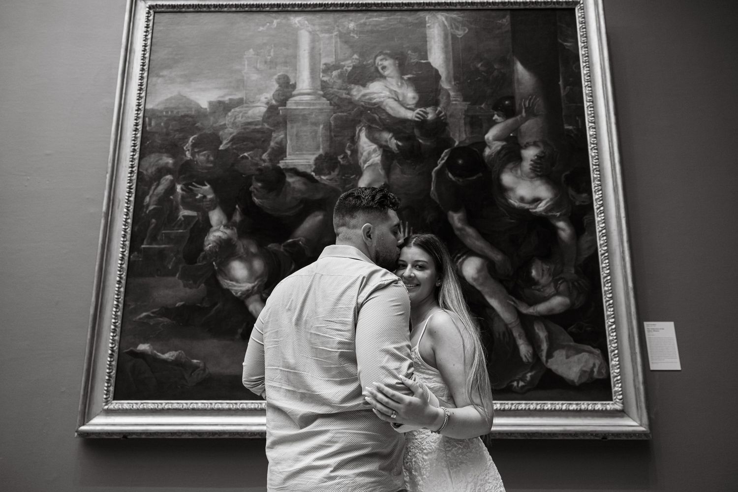 Couple embracing in black and white photo before a large classical painting in an art museum gallery.