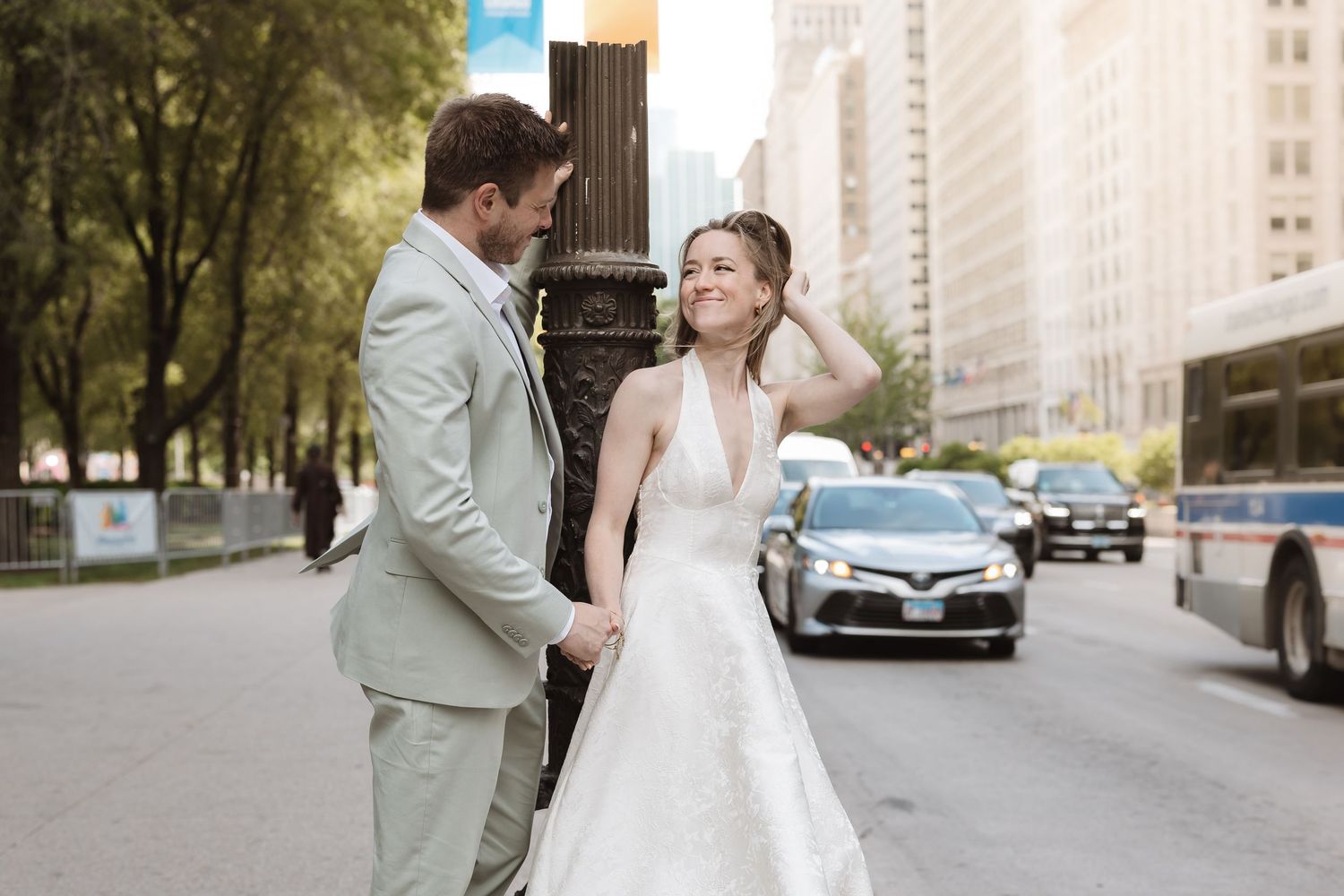 Bride and groom holding hands on a city street, smiling at each other in wedding attire with urban traffic behind them.