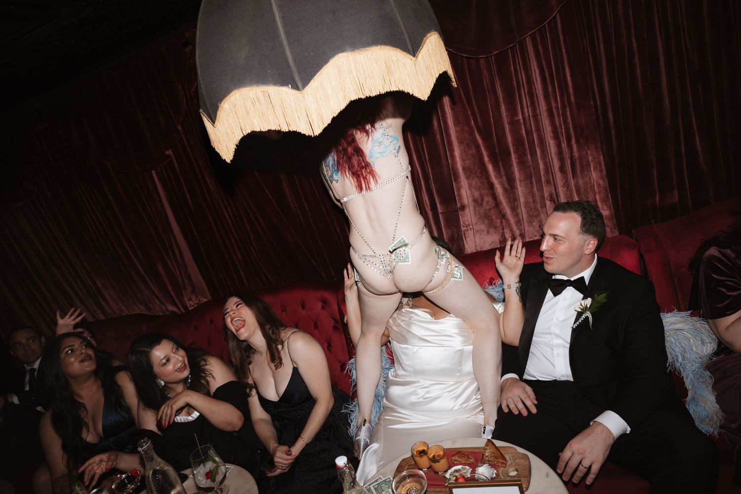 Bride flips dress up at wedding reception while guests laugh on red velvet sofa.