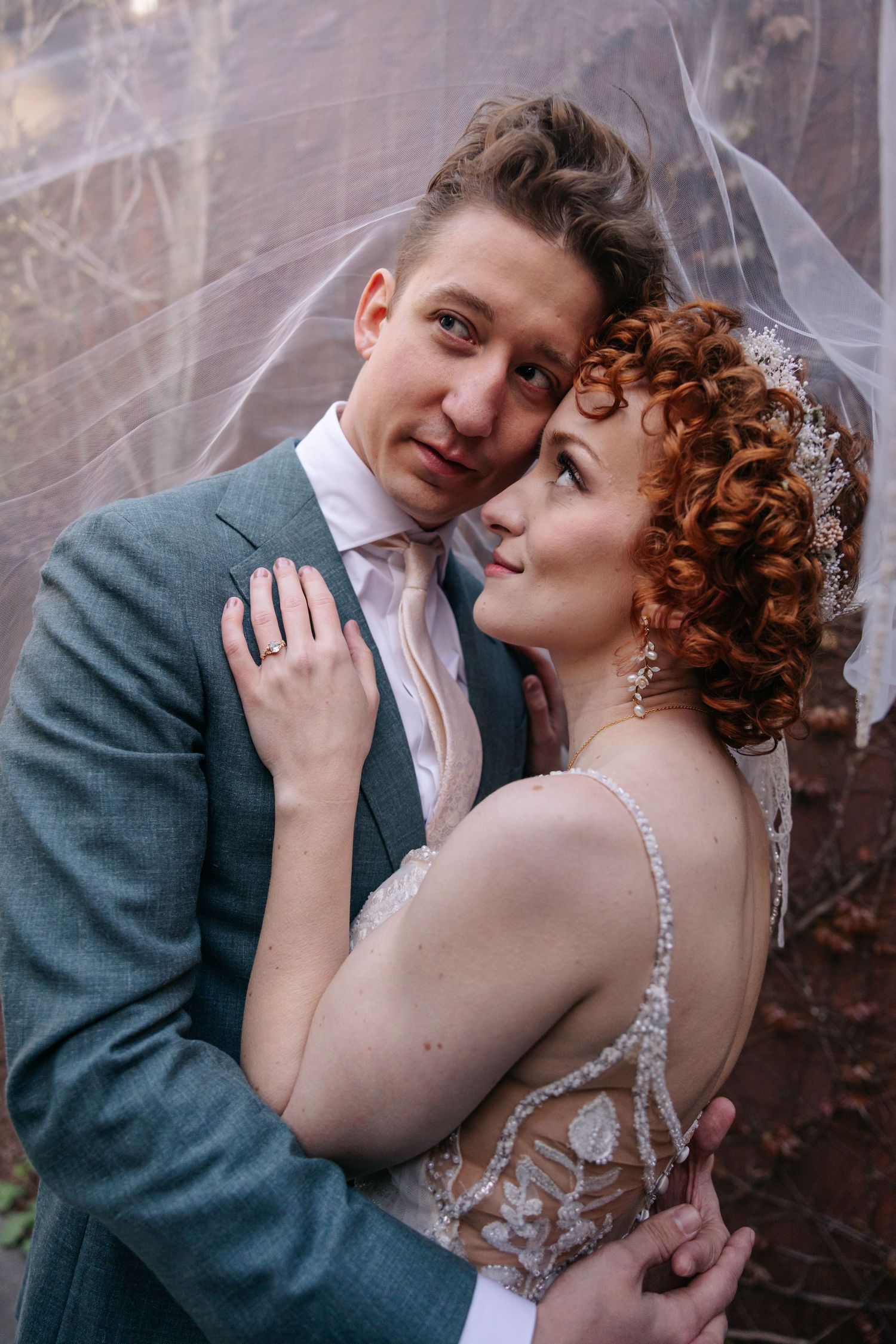 Bride with red curly hair and groom in teal suit posing under veil against autumn foliage backdrop.