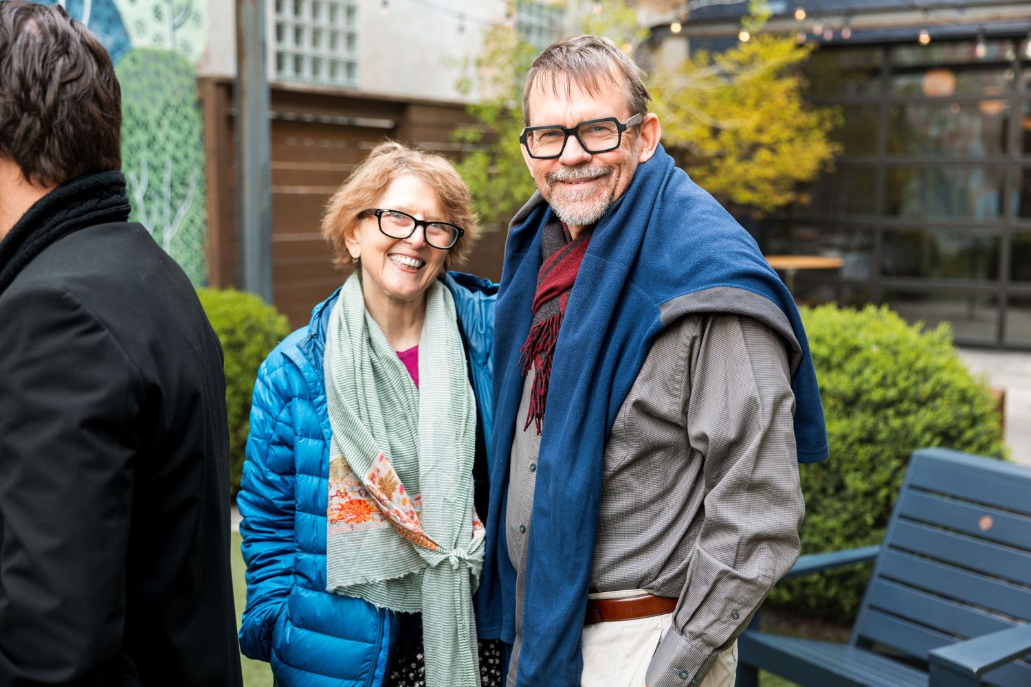 Smiling older couple wearing glasses and colorful scarves posing together outdoors at a social gathering.