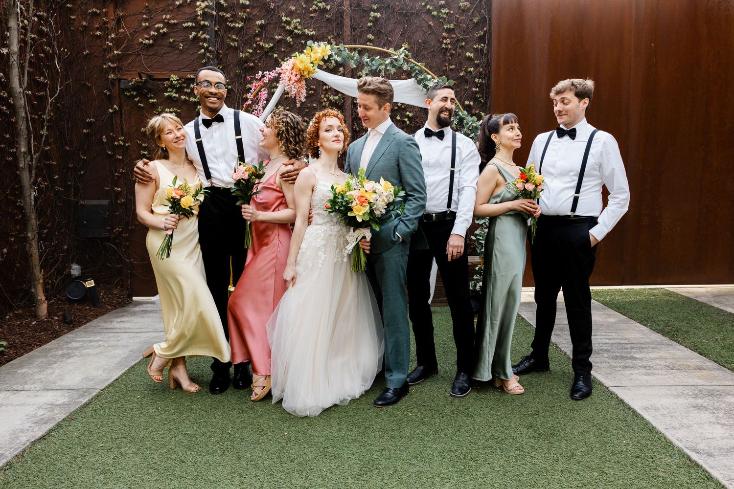Wedding party poses together in front of a floral hoop arch, bridesmaids in colorful gowns and groomsmen in suspenders.