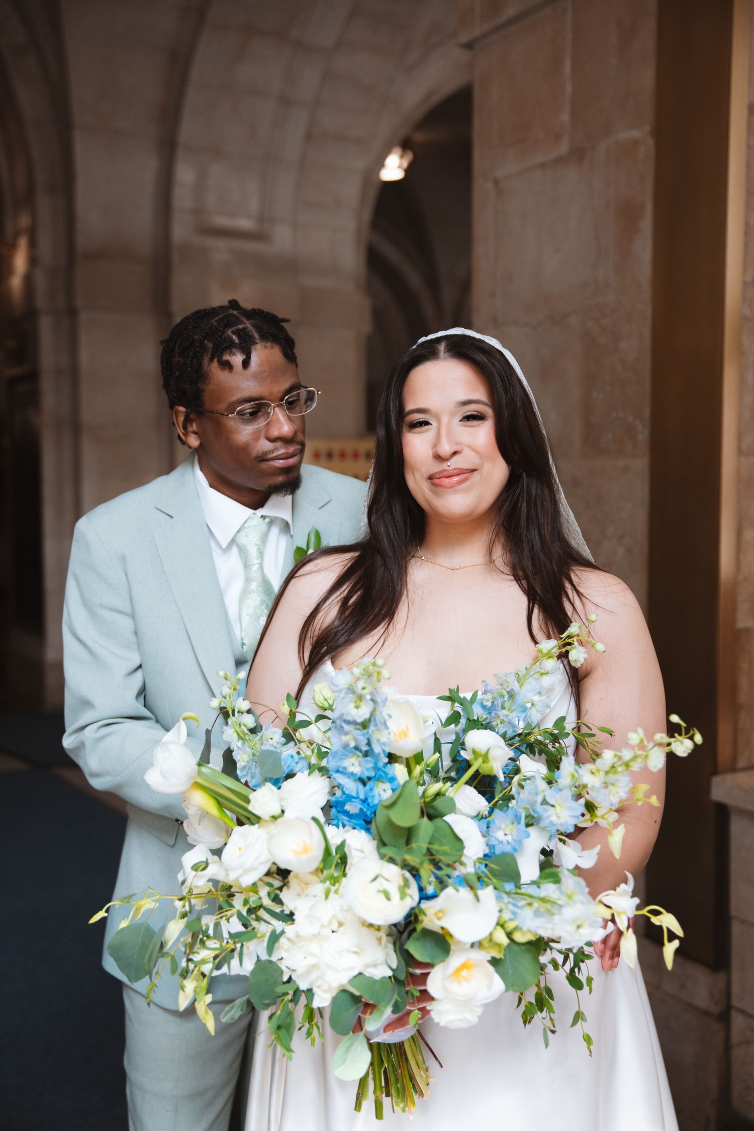 Interracial couple poses for wedding portrait, bride holding blue and white bouquet, groom in light gray suit.