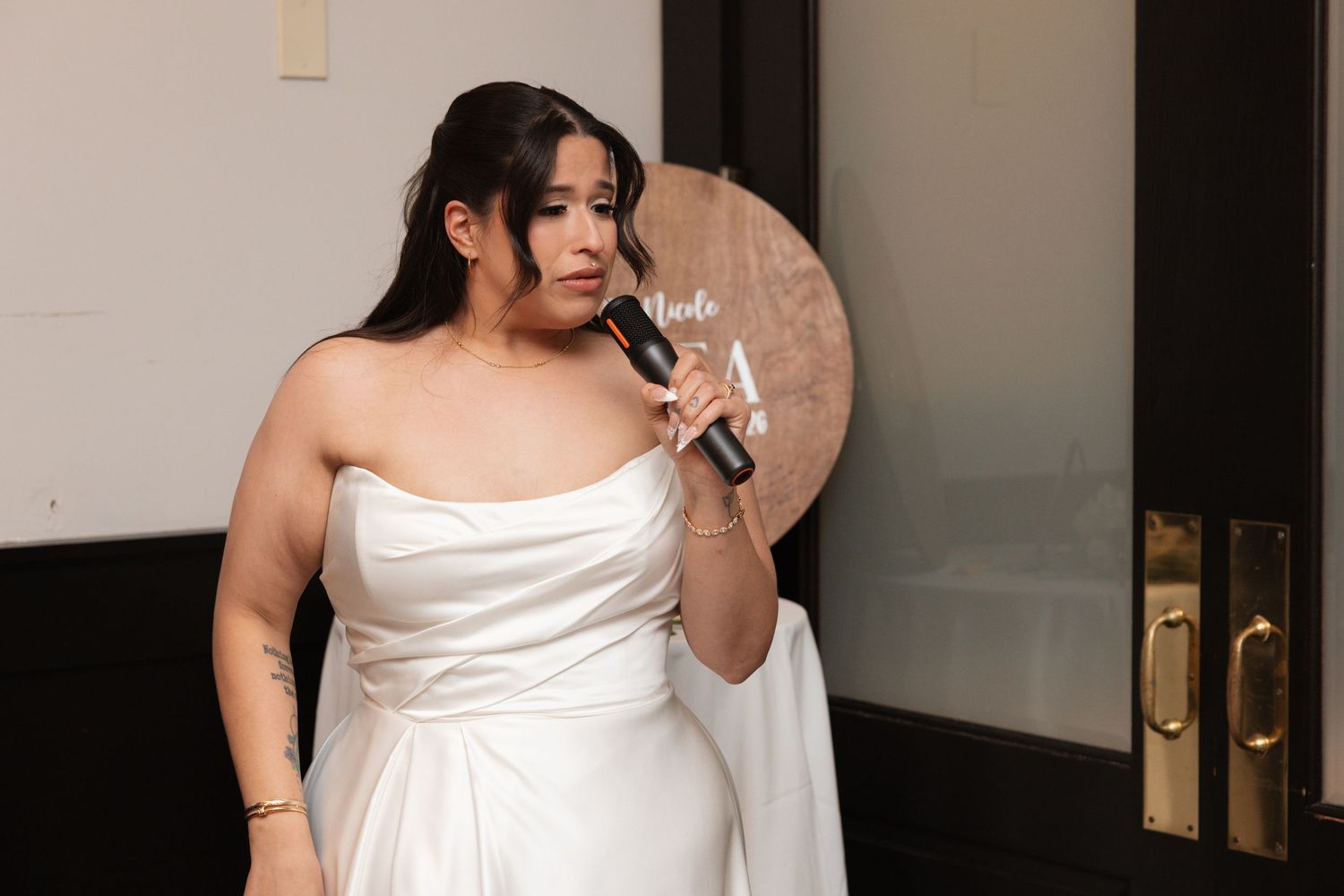 Bride in strapless white wedding gown speaking into microphone at reception.