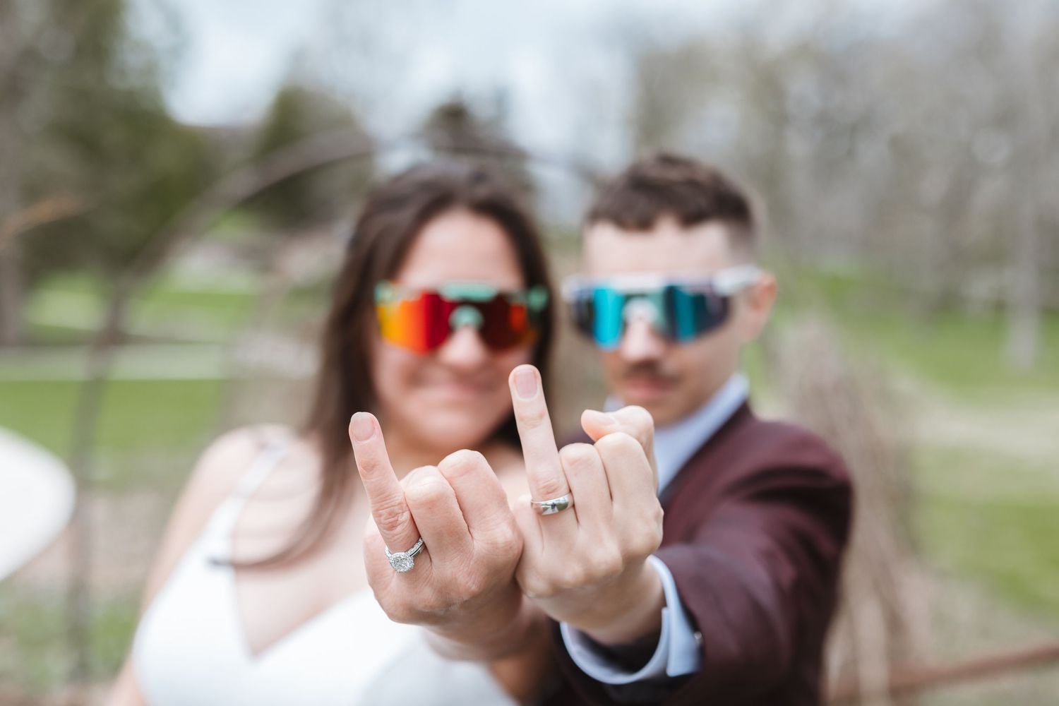Newlyweds in colorful sunglasses showing off wedding rings with rock on hand gesture outdoors.