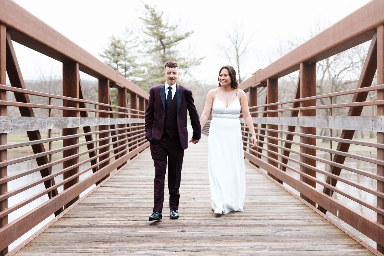 A bride and groom walk hand-in-hand on a wooden bridge, smiling, dressed in a white gown and dark suit.