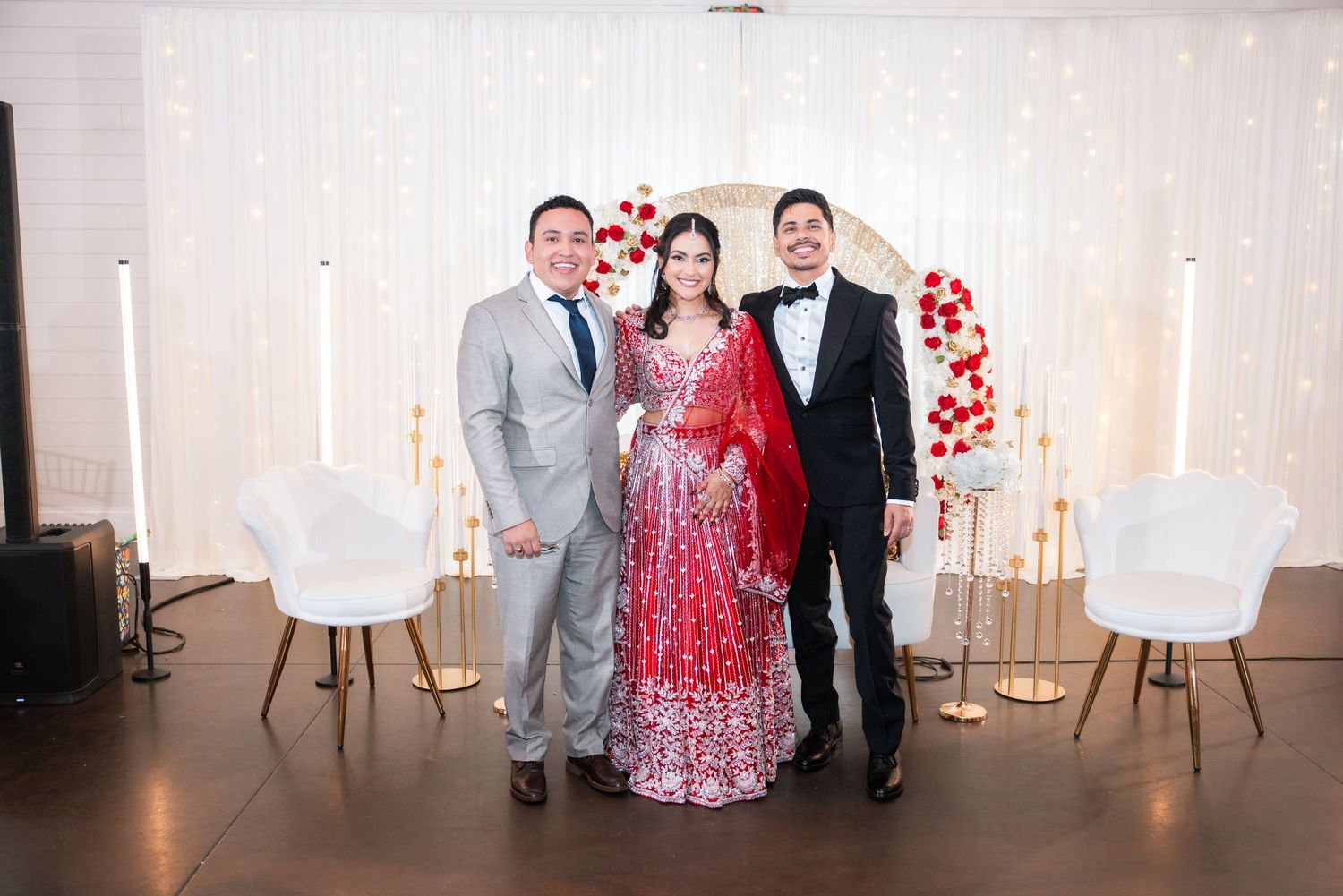 Bride in red lehenga and groom in tuxedo pose with a guest at a fairy-lit wedding reception.