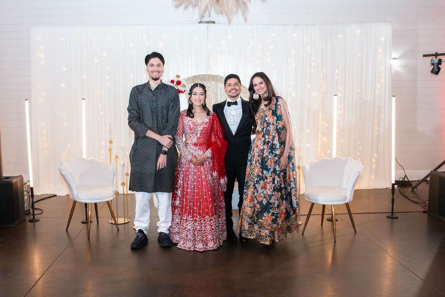 Four people posing together at an Indian wedding celebration, bride wearing red lehenga with floral white backdrop.