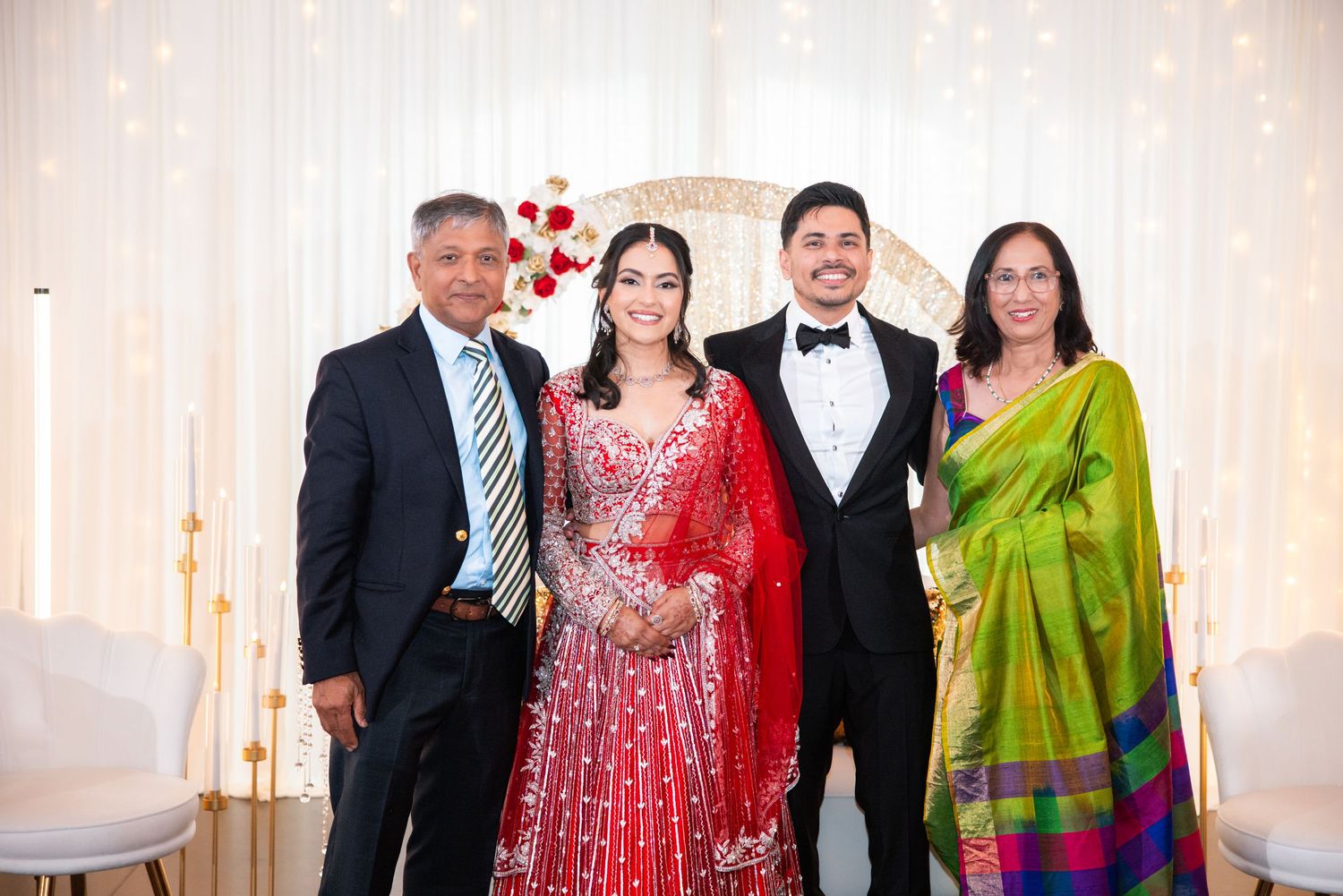 Indian wedding family portrait with bride in red lehenga and groom in tuxedo with parents.